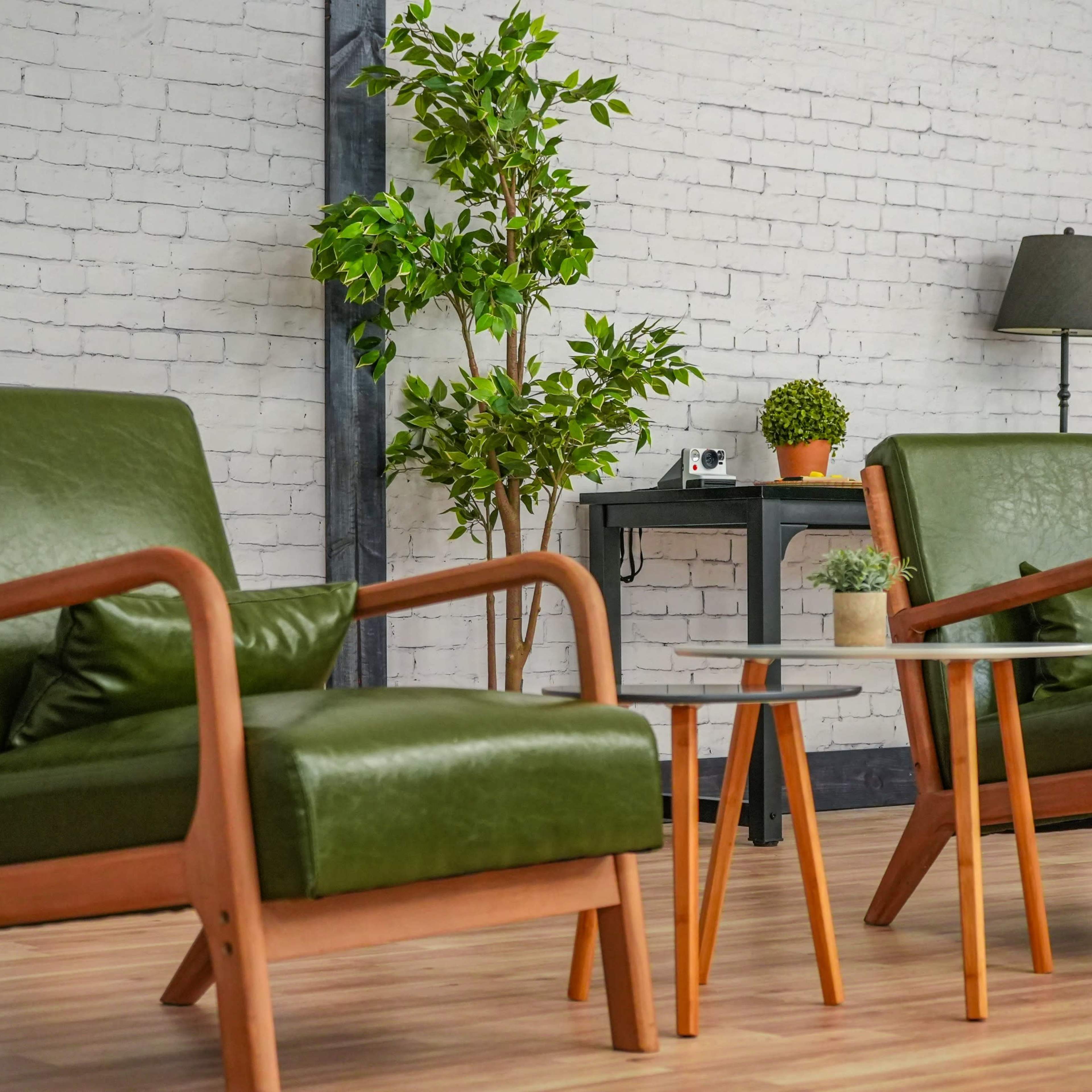 Two green armchairs with wooden frames positioned around a small round table in a room with a light brick wall and potted plants.