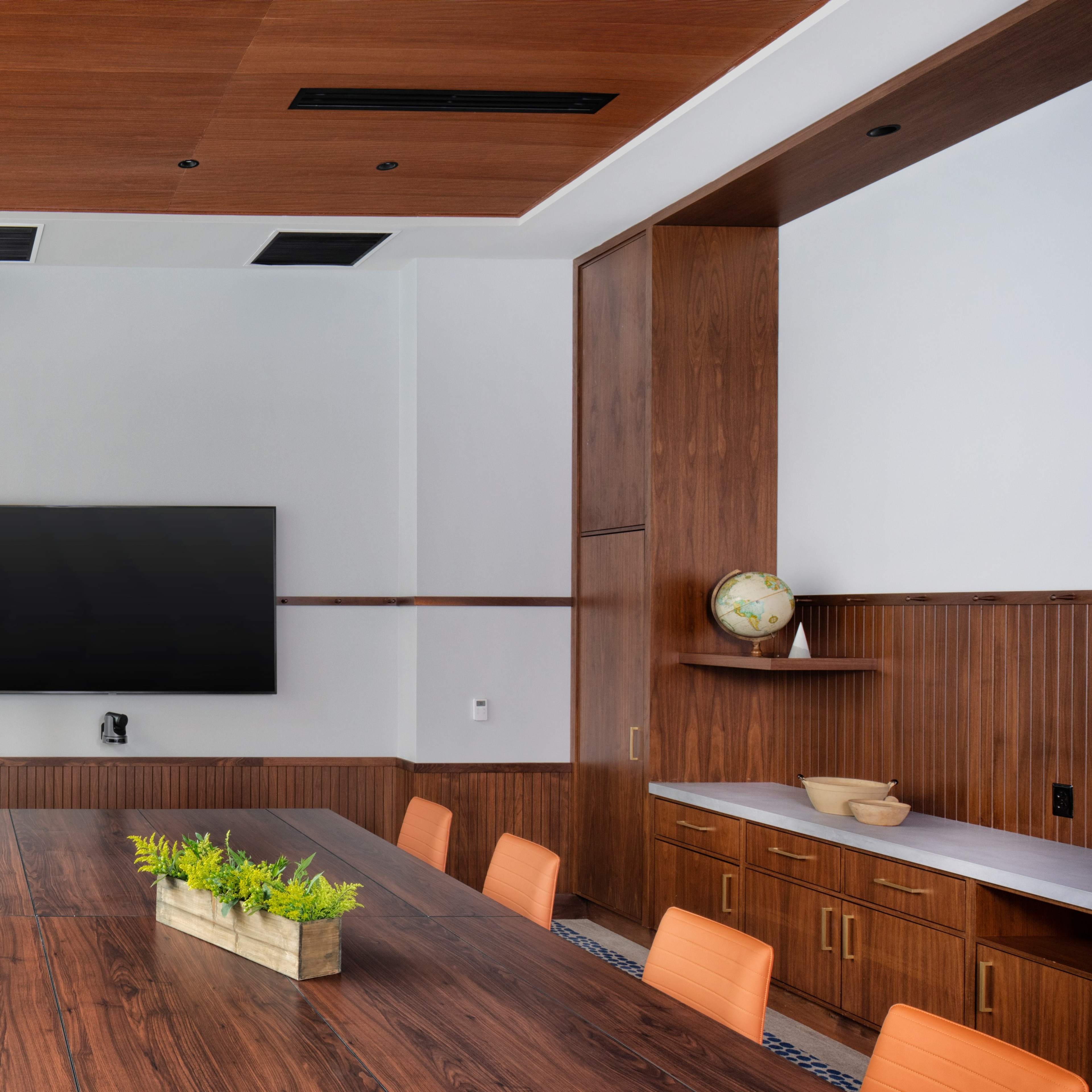 A modern conference room features a long wooden table, a flat-screen TV on the wall, and a decorative planter at the center.