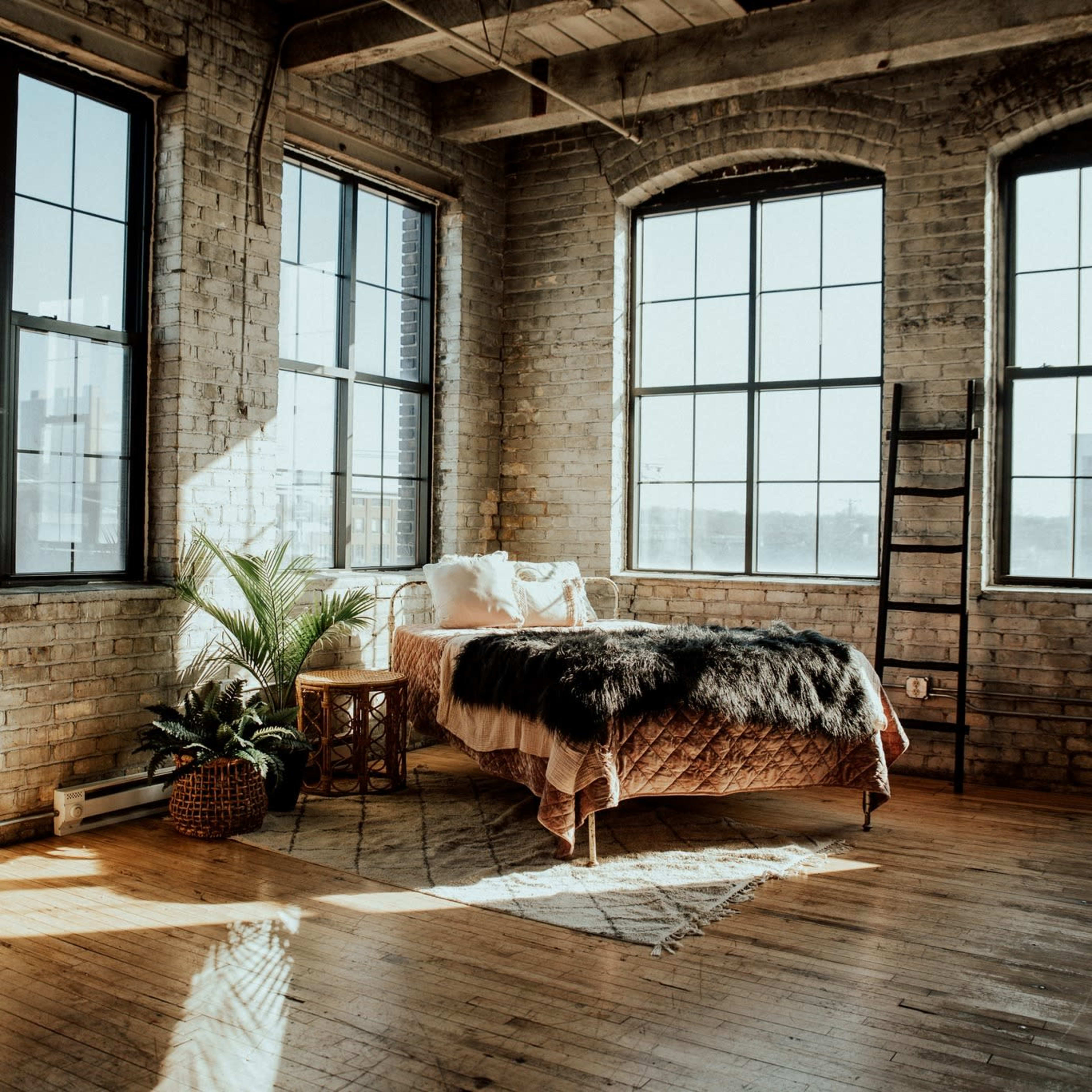 A sunlit bedroom features a bed with a fur blanket, large windows, and a ladder against a brick wall.