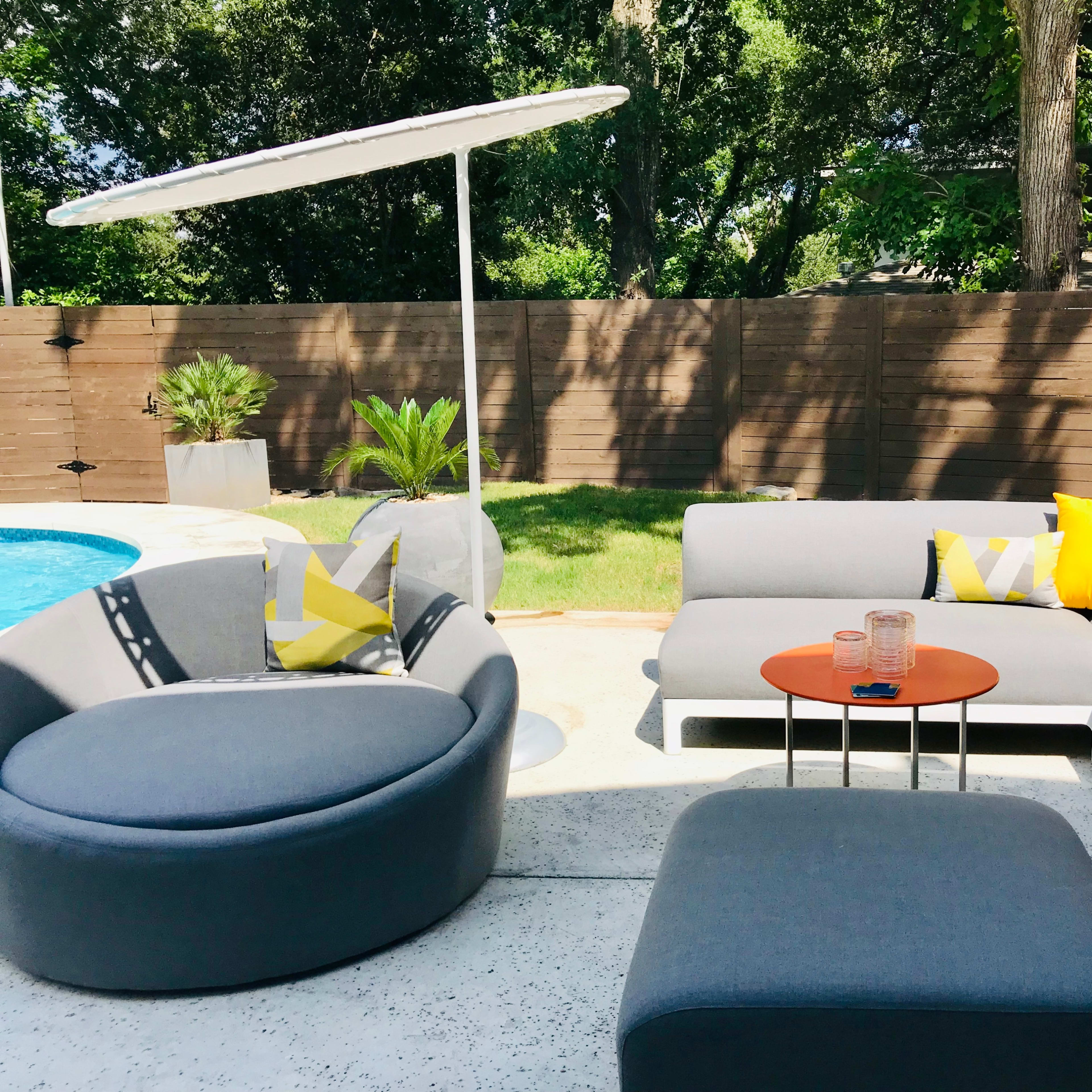 The scene showcases a modern outdoor patio with gray furniture, a small orange table, and a pool surrounded by lush greenery.