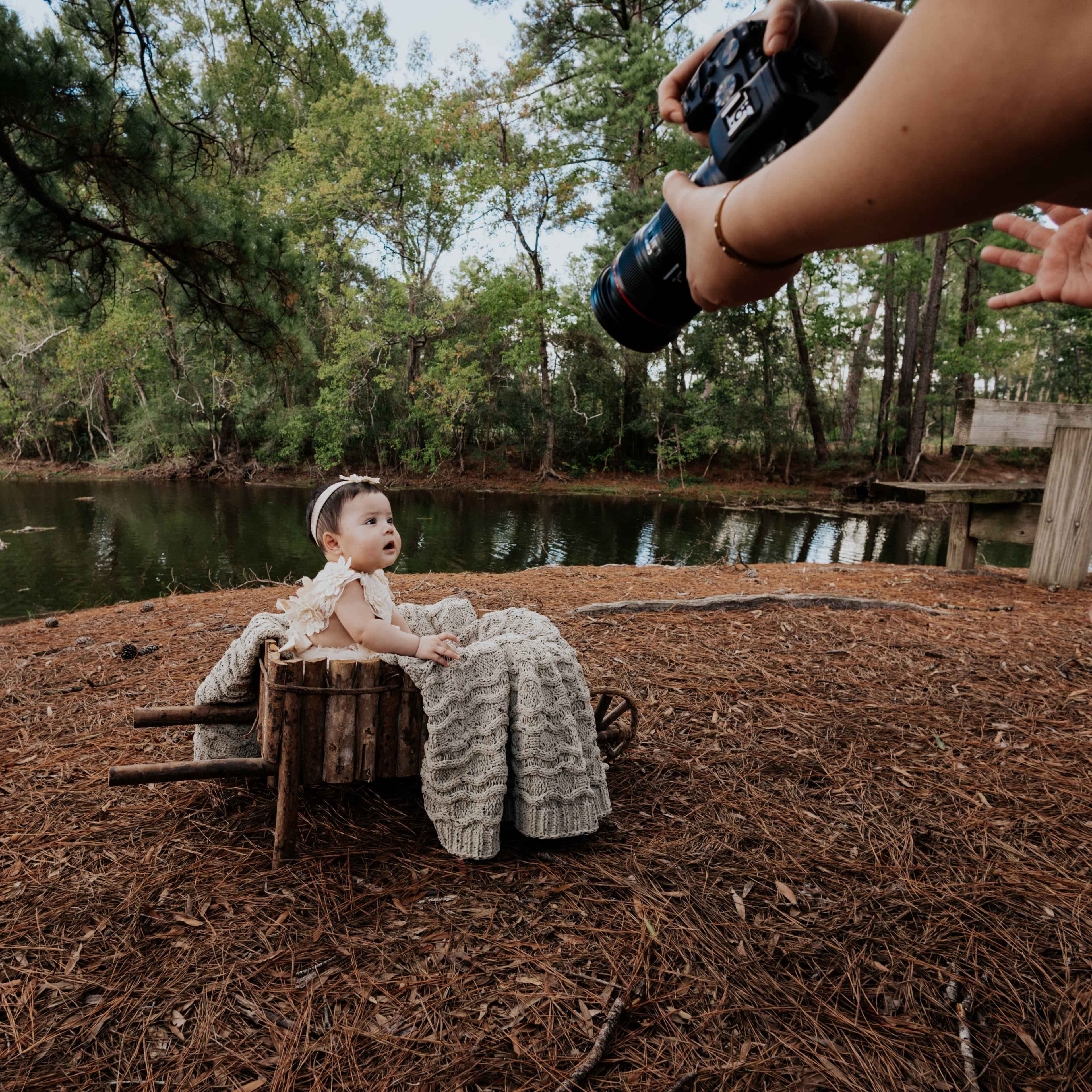 A baby sits in a wooden cart on a bed of pine needles by a pond, while a person uses a camera to take photos.