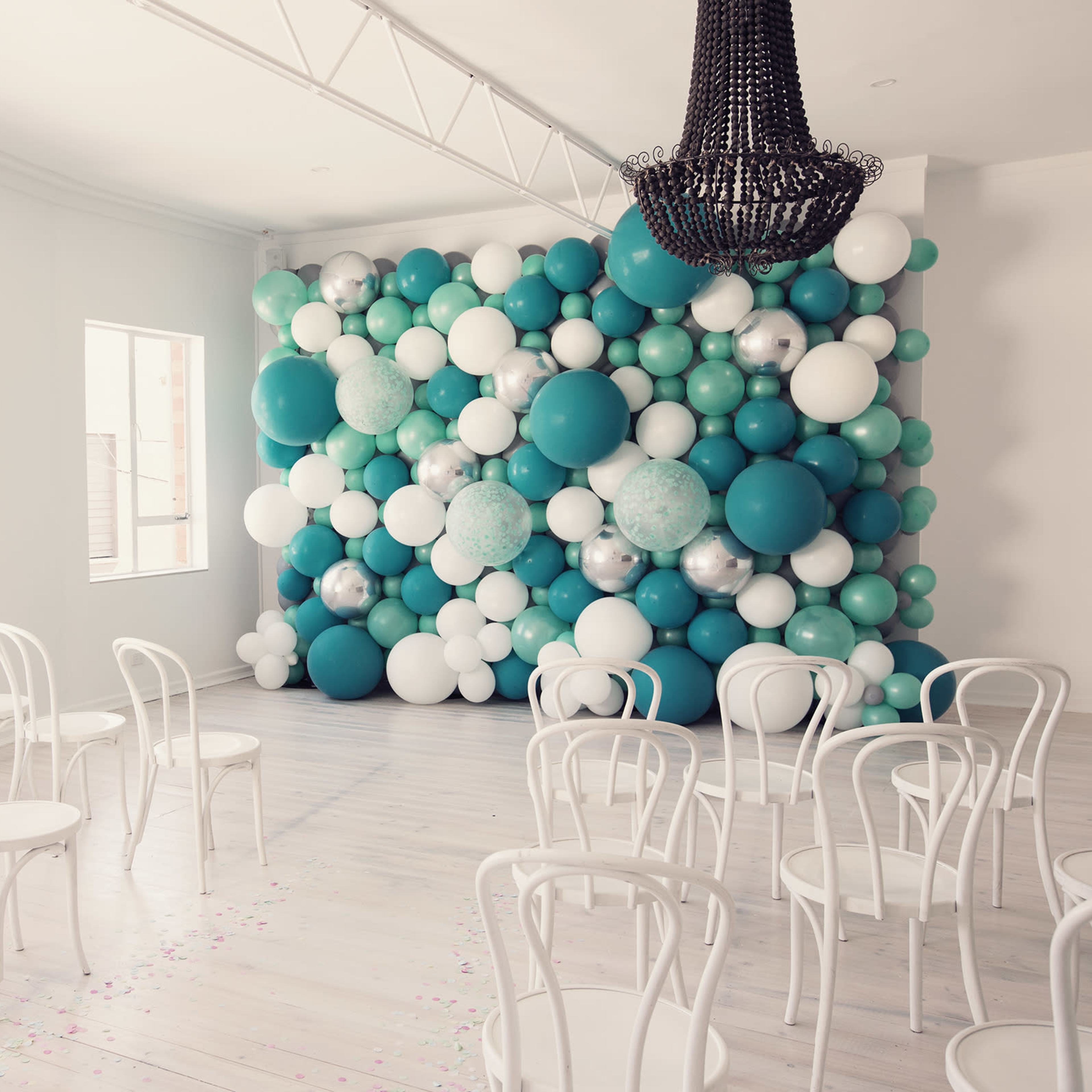 A room with a decorative wall of balloons in shades of teal and white, accompanied by several white chairs facing the wall.