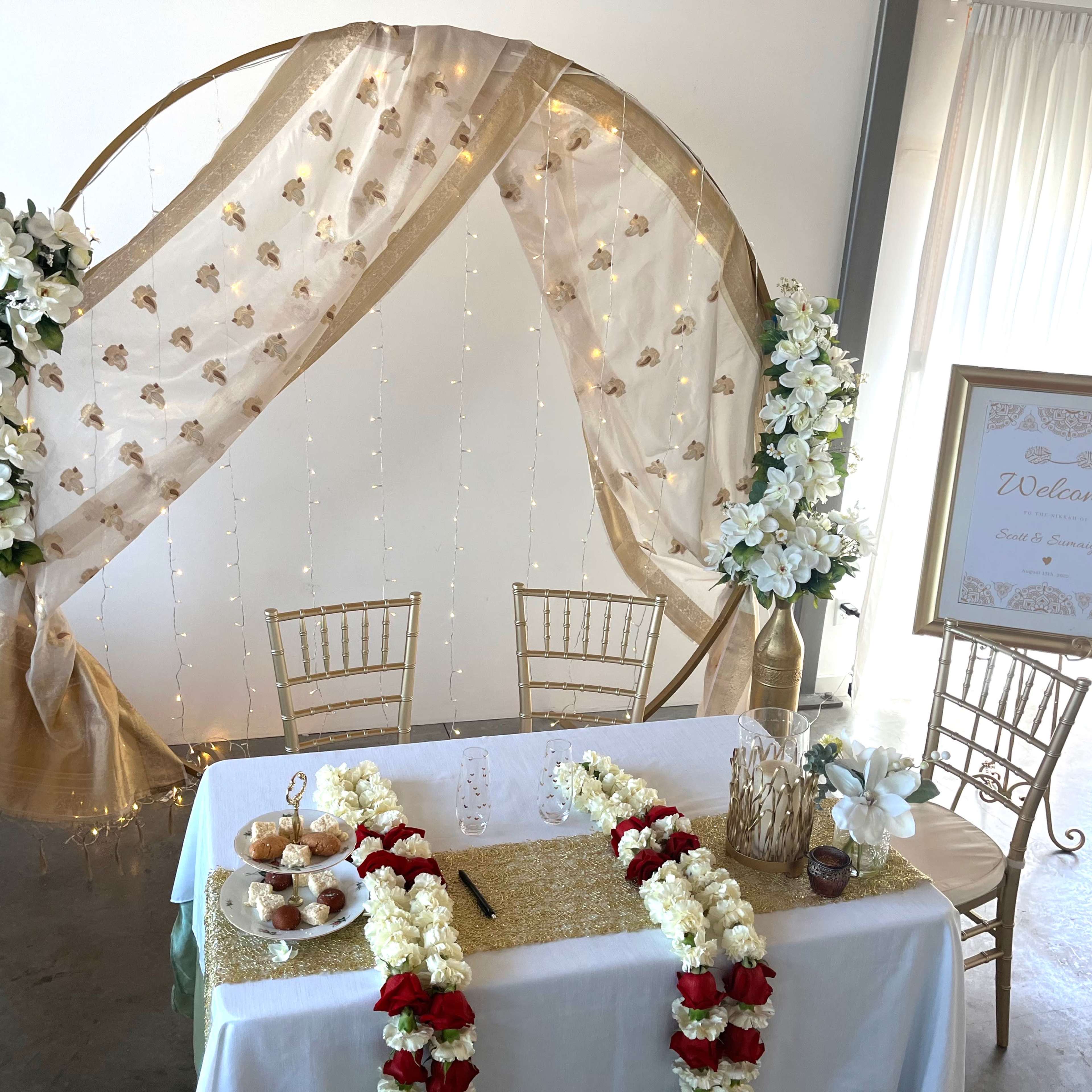 A decorative event setup features a gold circular backdrop draped with sheer fabric and adorned with white flowers and fairy lights, accompanied by a welcome sign and a table with a floral arrangement and refreshments.