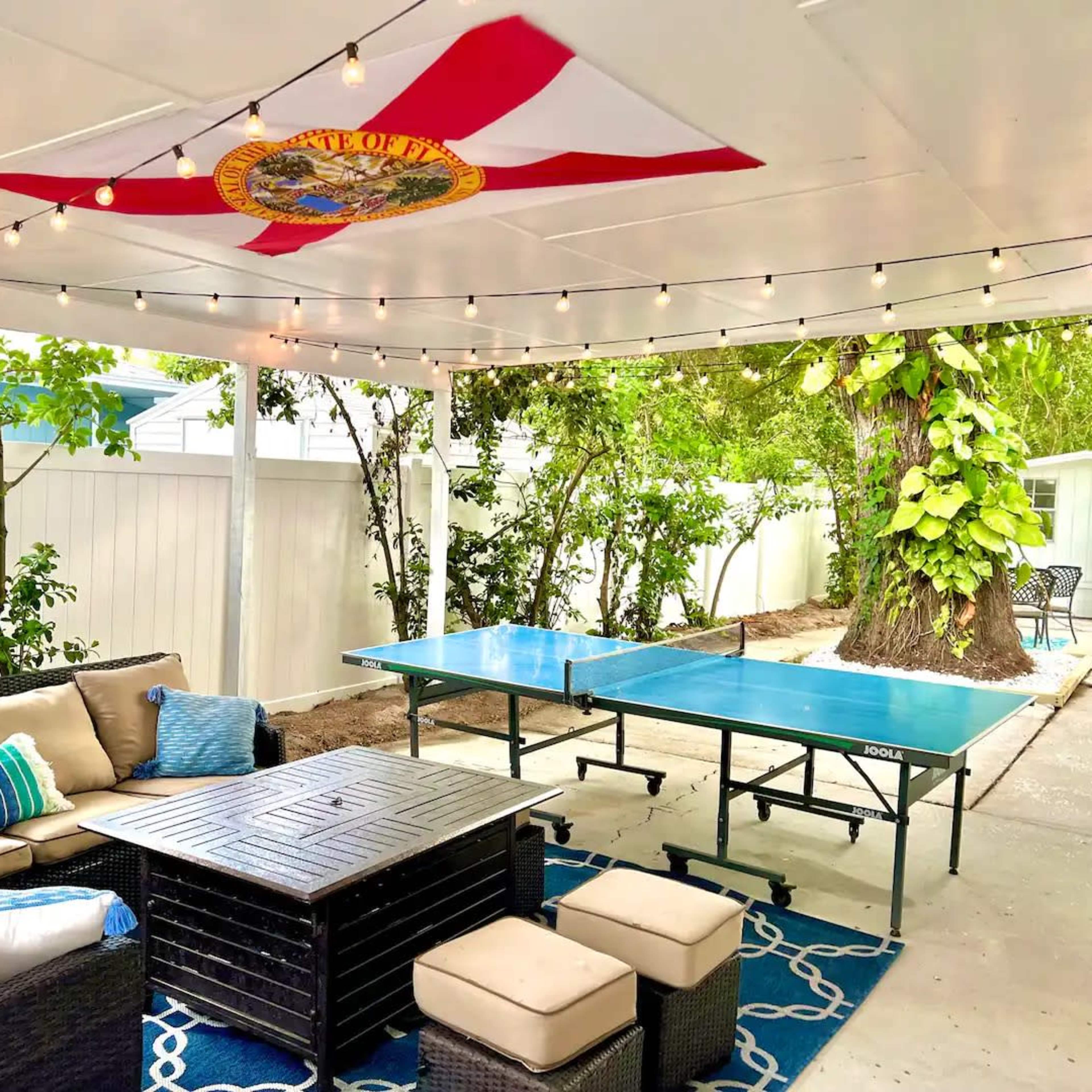 A covered outdoor patio features a seating area with a sectional sofa and coffee table, alongside a ping pong table under string lights and a flag hanging from the ceiling.