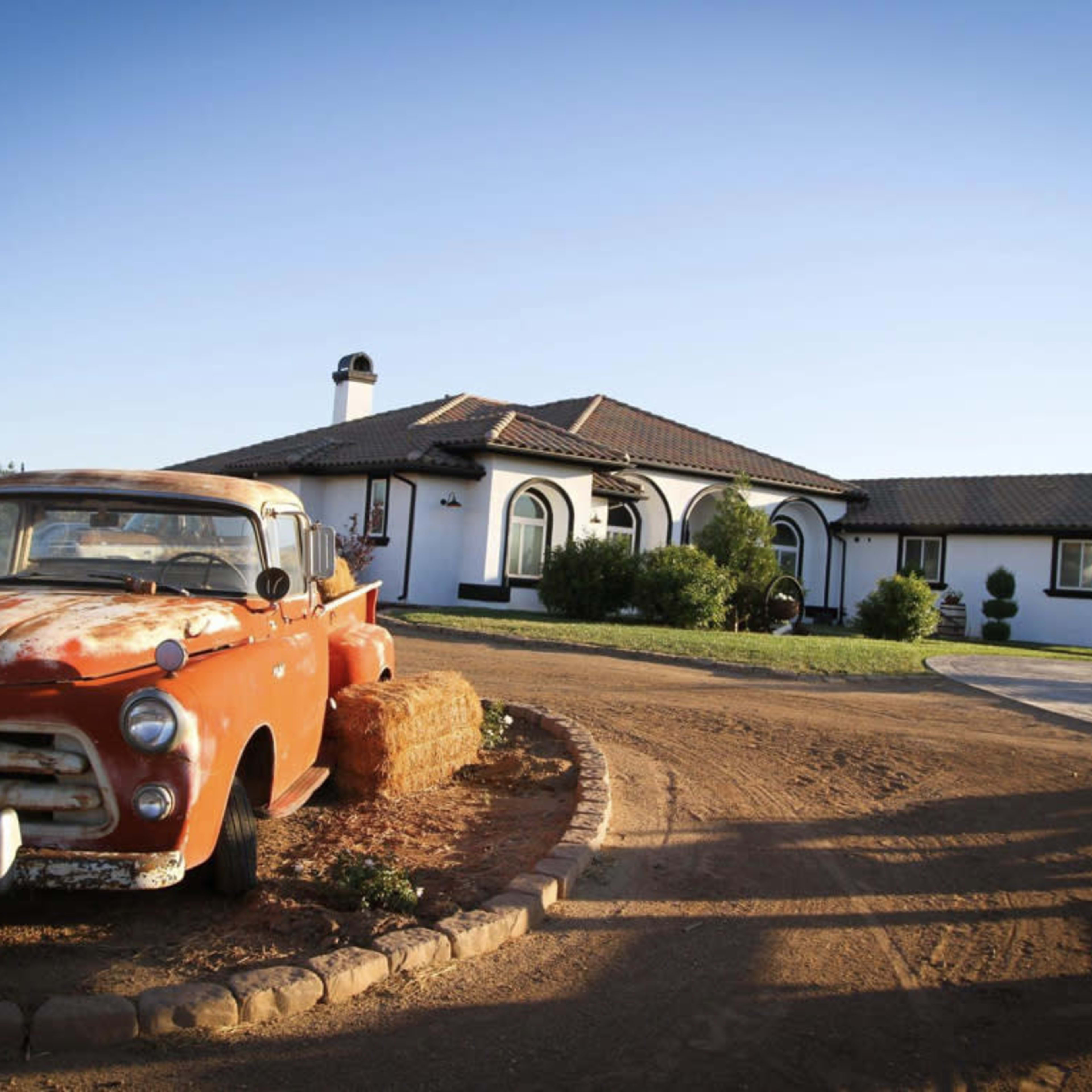 An old orange truck is parked beside a winding driveway leading to a modern white house with a tiled roof.