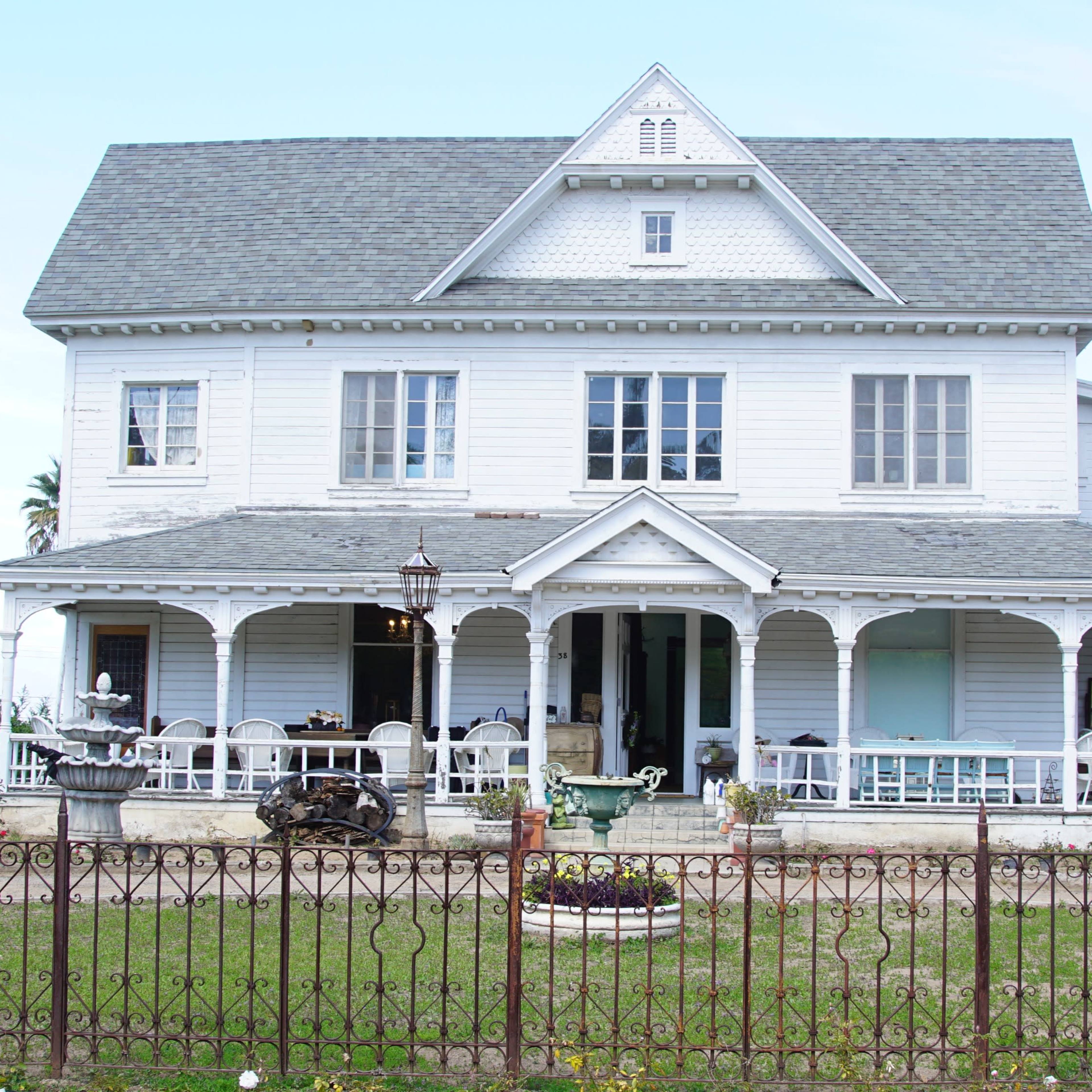 A large, two-story white house with a decorative porch and a well-maintained yard is surrounded by a wrought-iron fence.