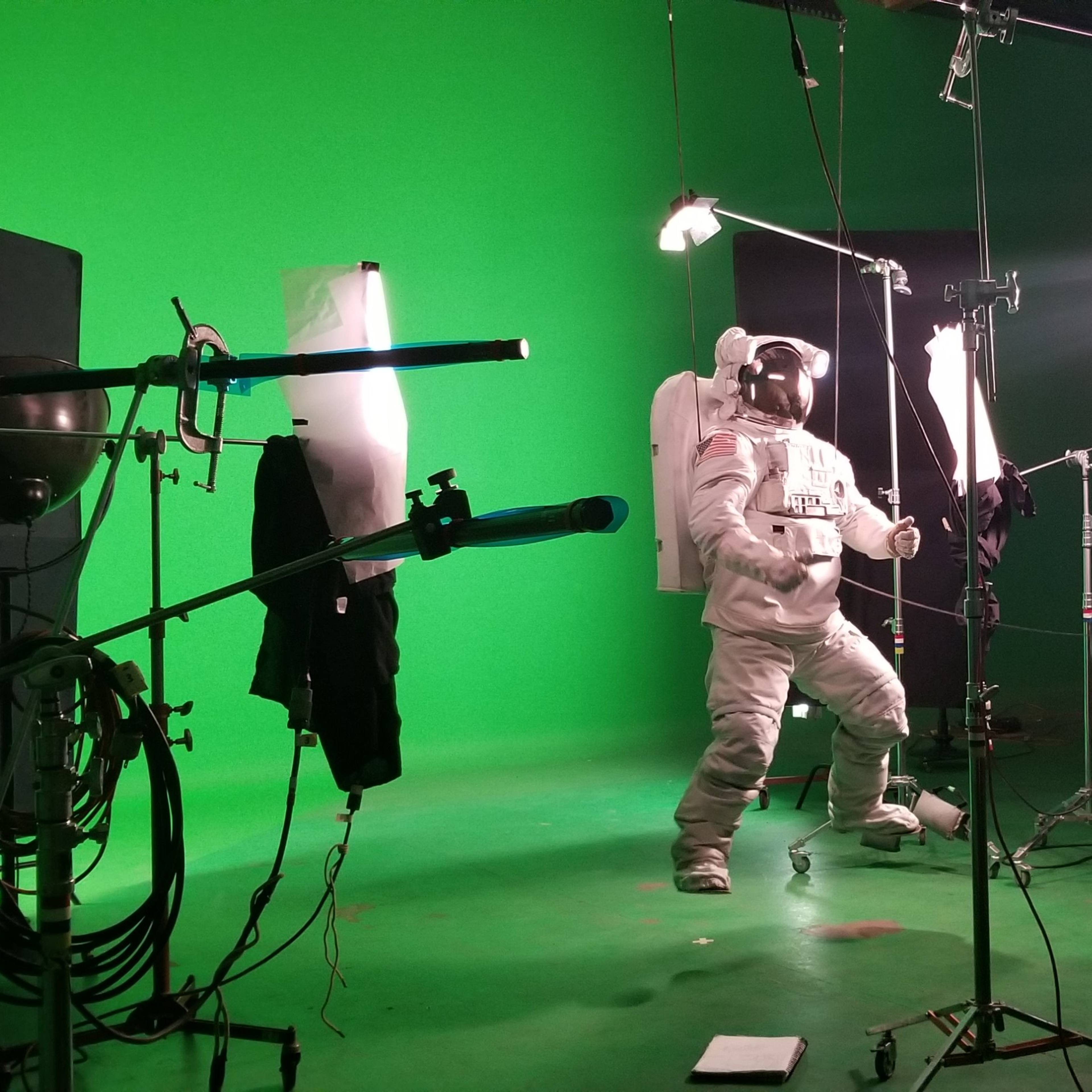 A person in a space suit stands in front of a green screen while filming equipment and lights are positioned around them.
