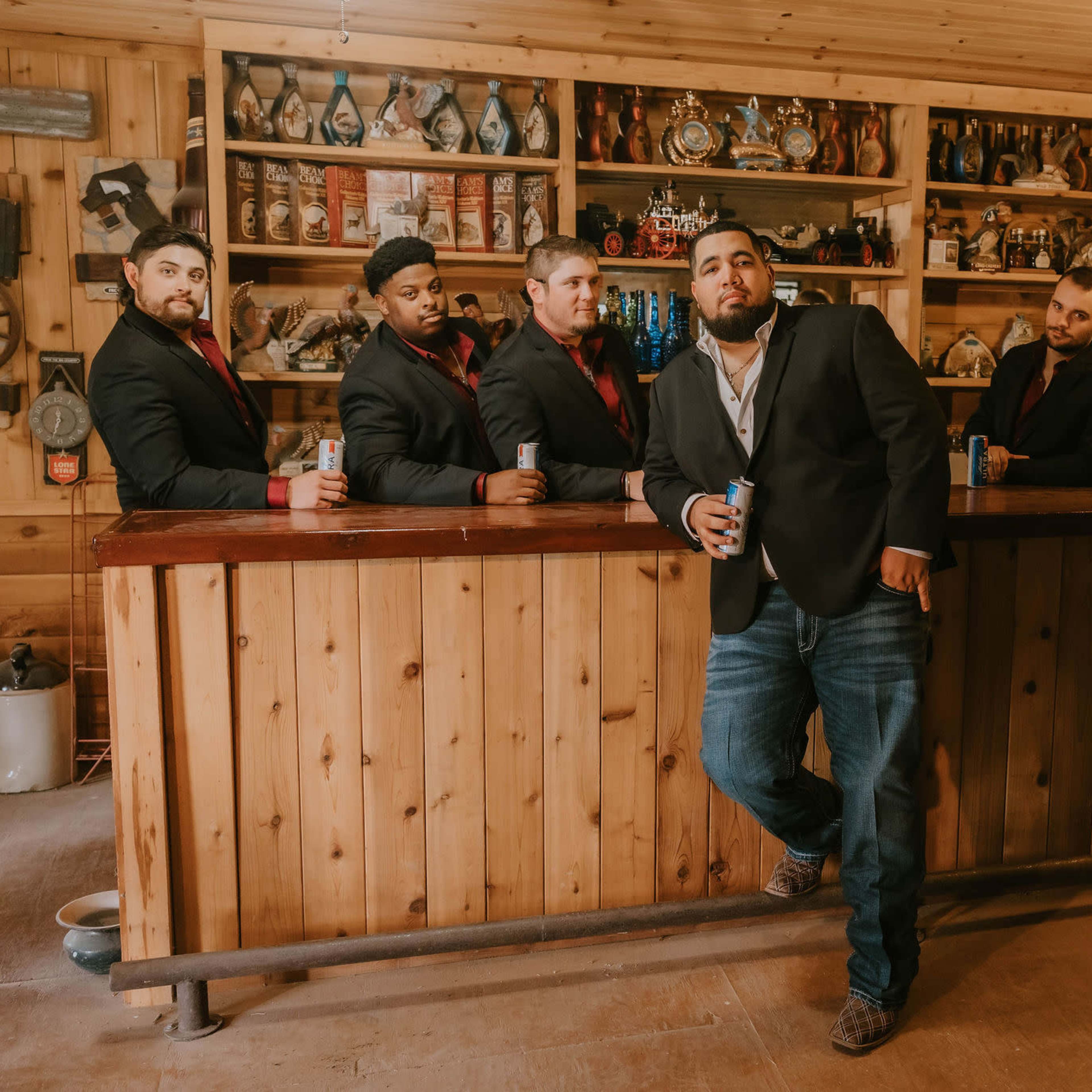Seven men in tuxedos stand behind a wooden bar, while one man in a black jacket and jeans poses in front, holding a can.