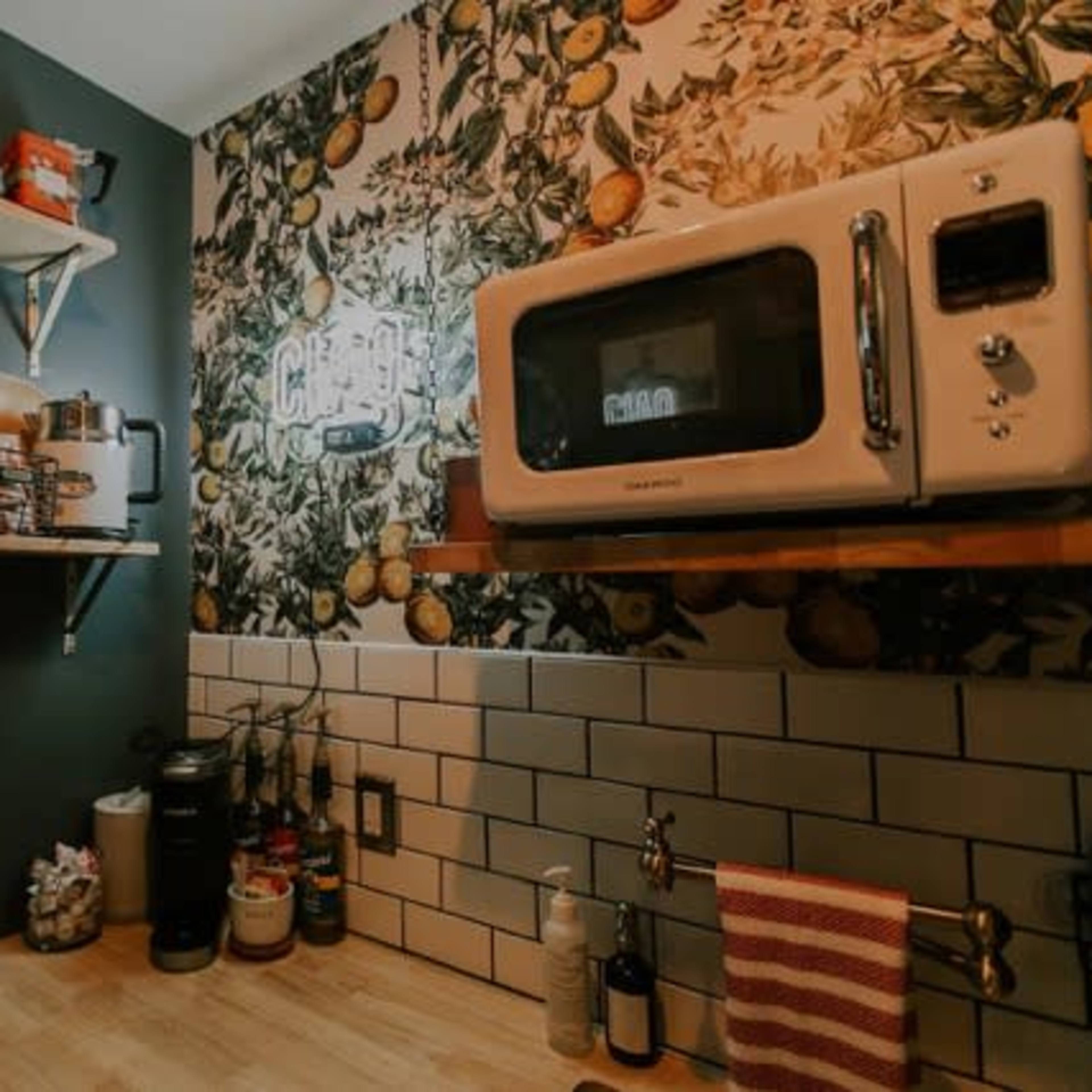 A small kitchen area features a microwave, open shelves with various dishes and ingredients, and a wall adorned with a botanical wallpaper.