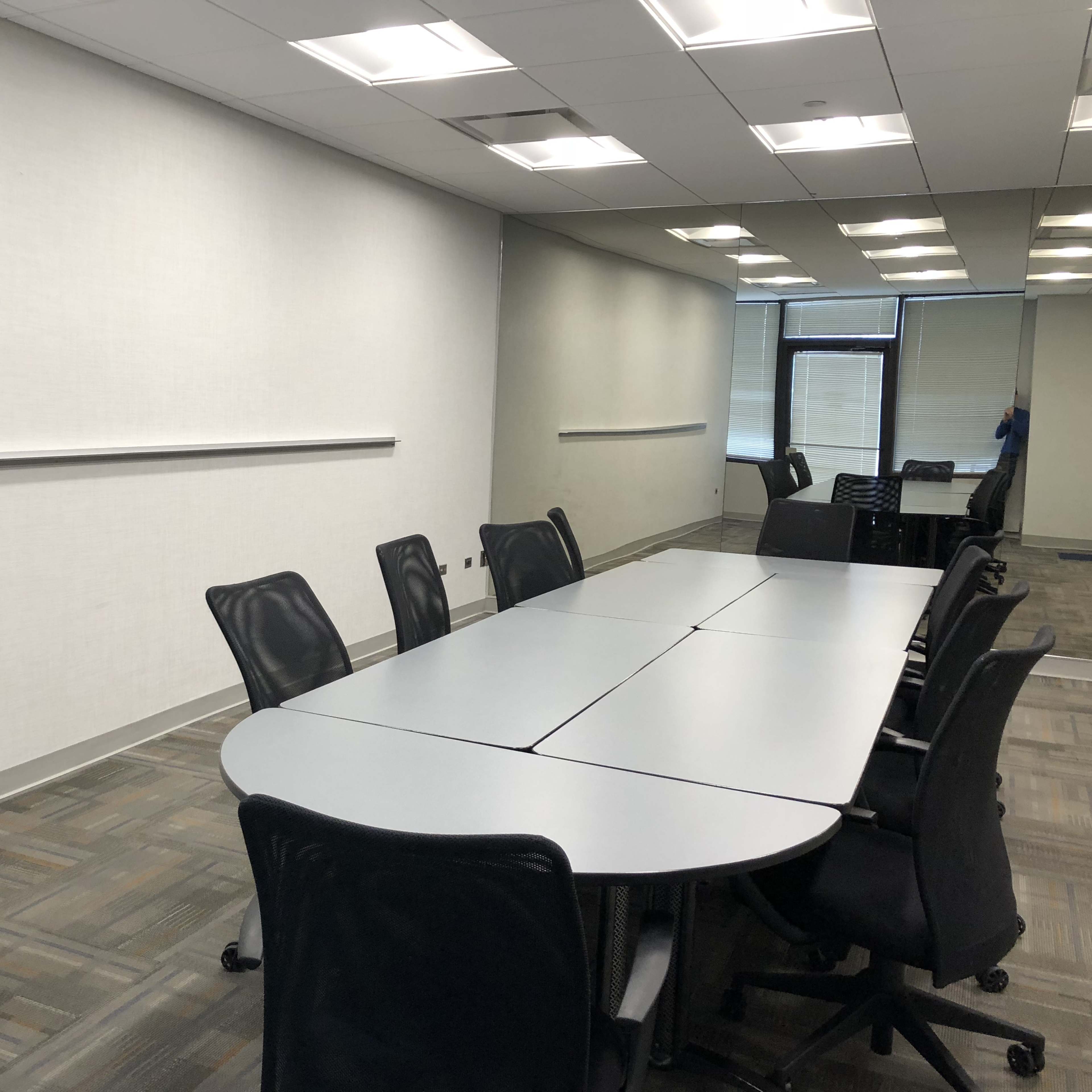 A large, rectangular conference table with black mesh chairs is positioned in a brightly lit meeting room with plain white walls.