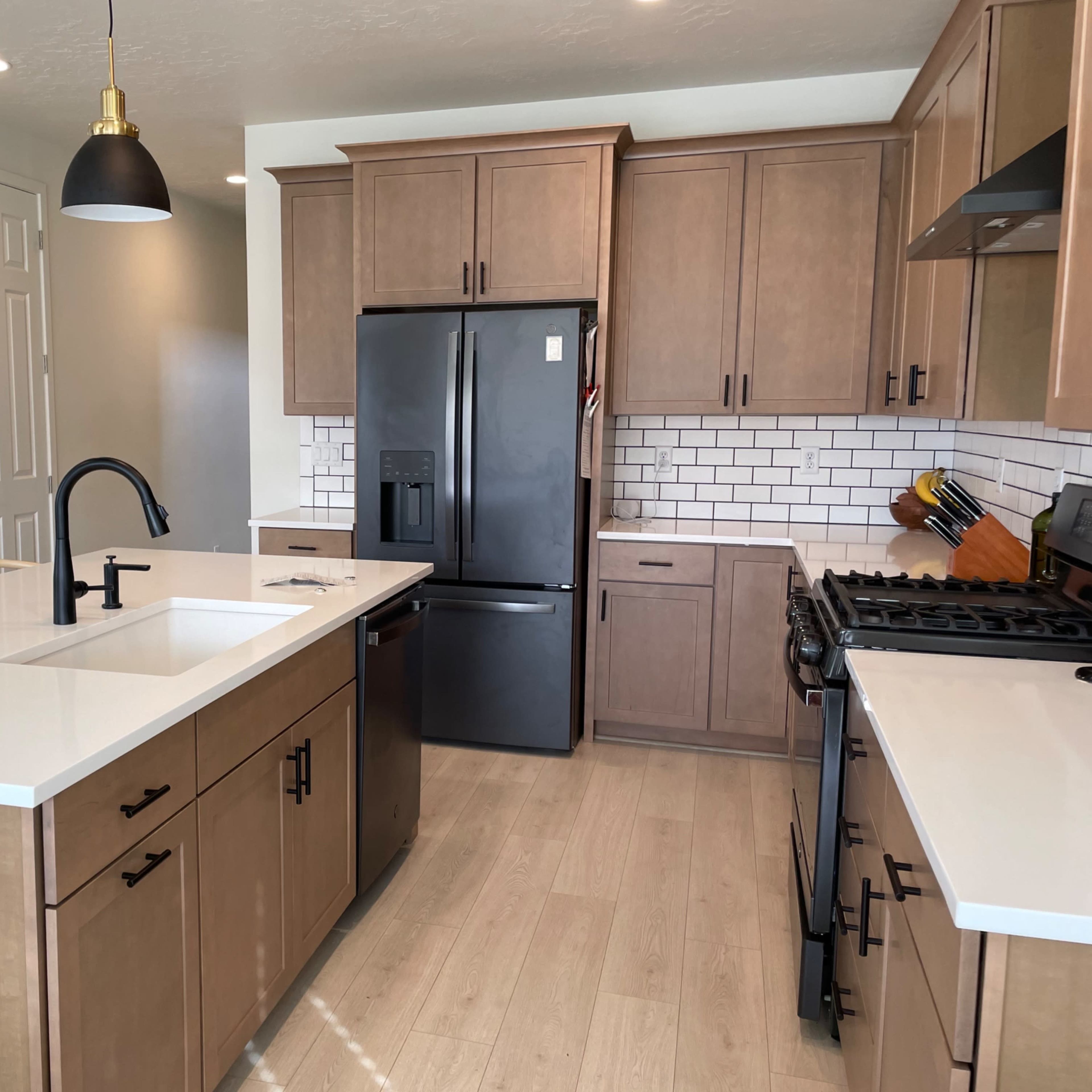 A modern kitchen featuring wooden cabinets, a black refrigerator, a white countertop, and a sink island with black pendant lights overhead.