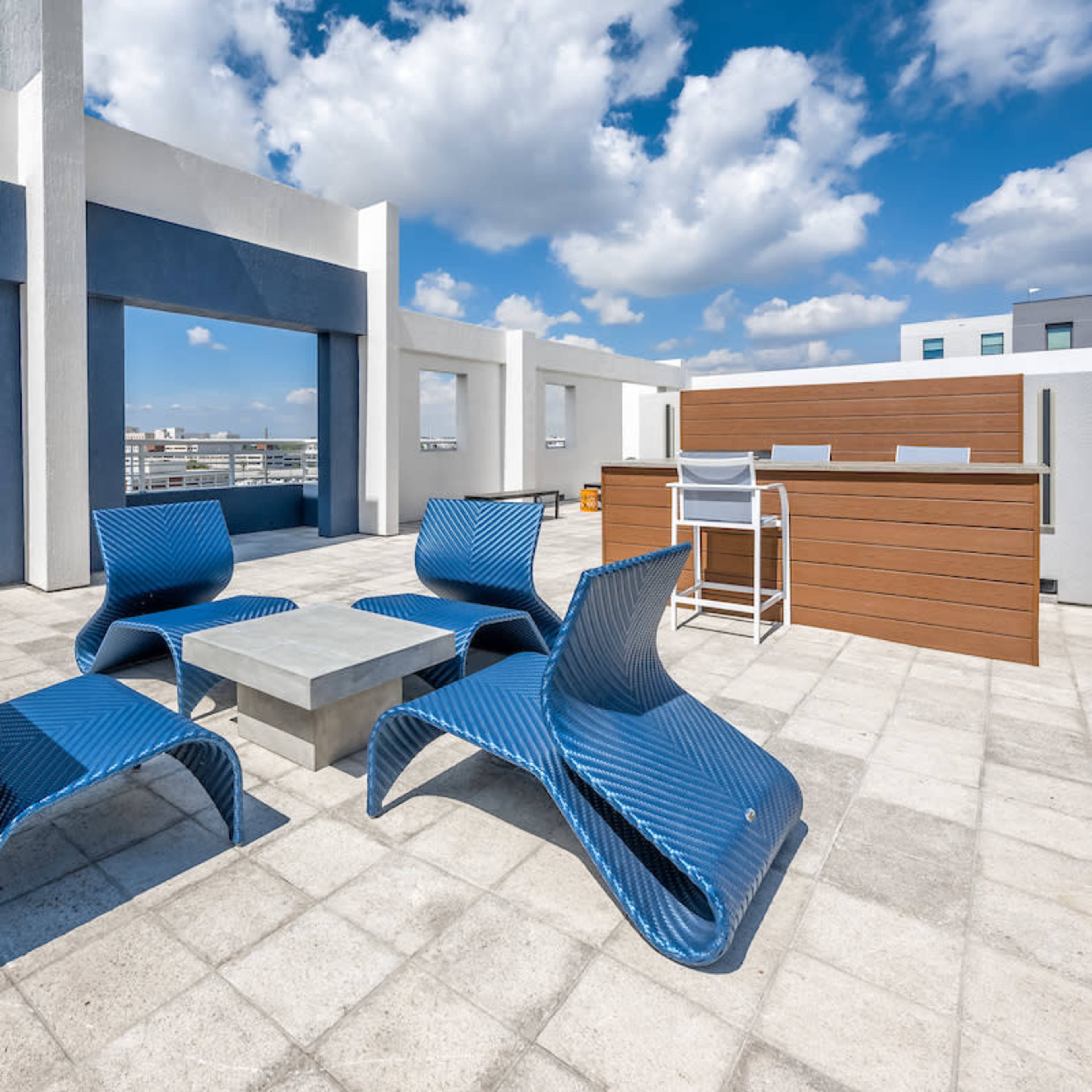A rooftop terrace featuring modern blue lounge chairs arranged around a concrete table, with a wooden bar area in the background and a clear blue sky above.