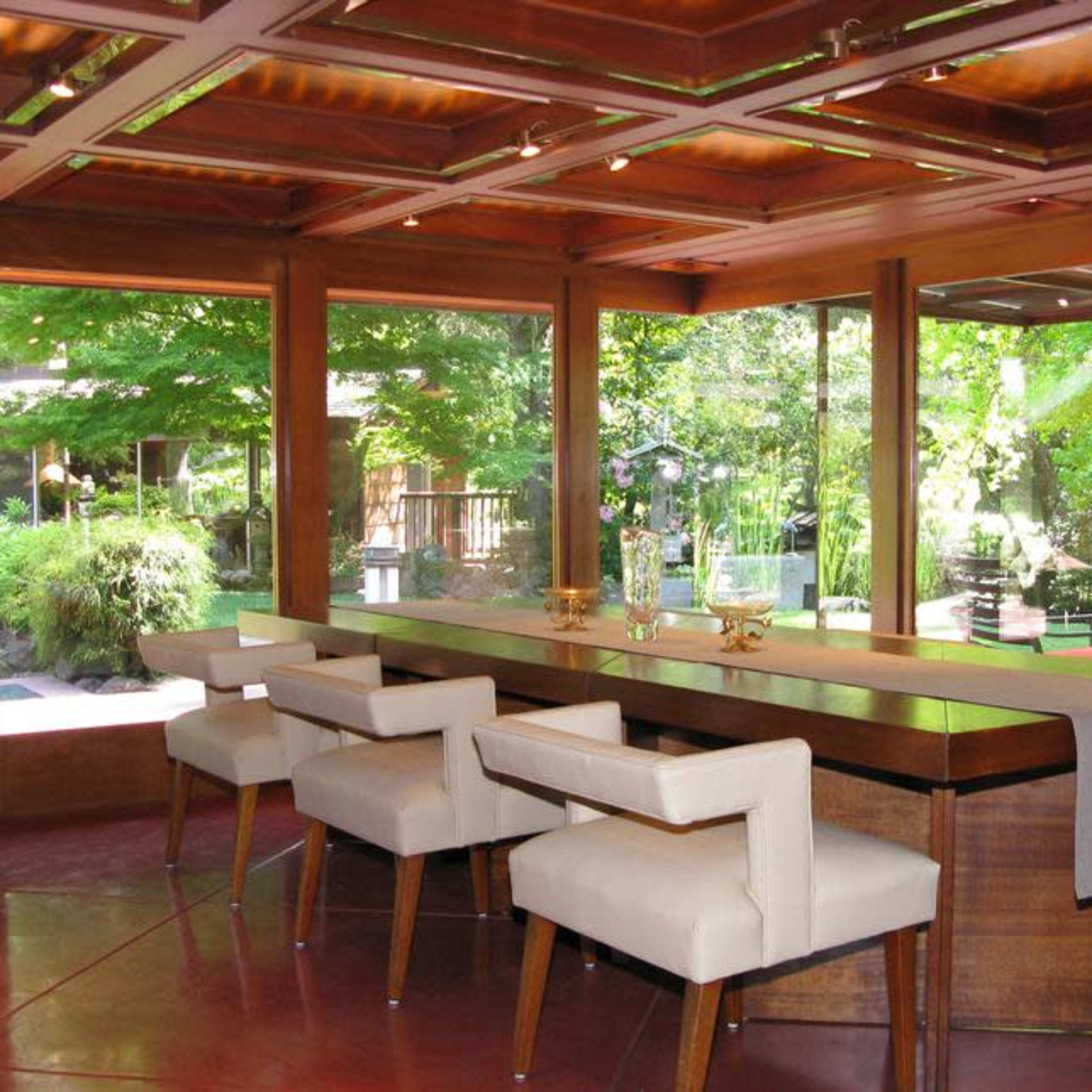 A dining area with a large wooden table surrounded by four white upholstered chairs, featuring large windows that provide views of a lush outdoor garden.