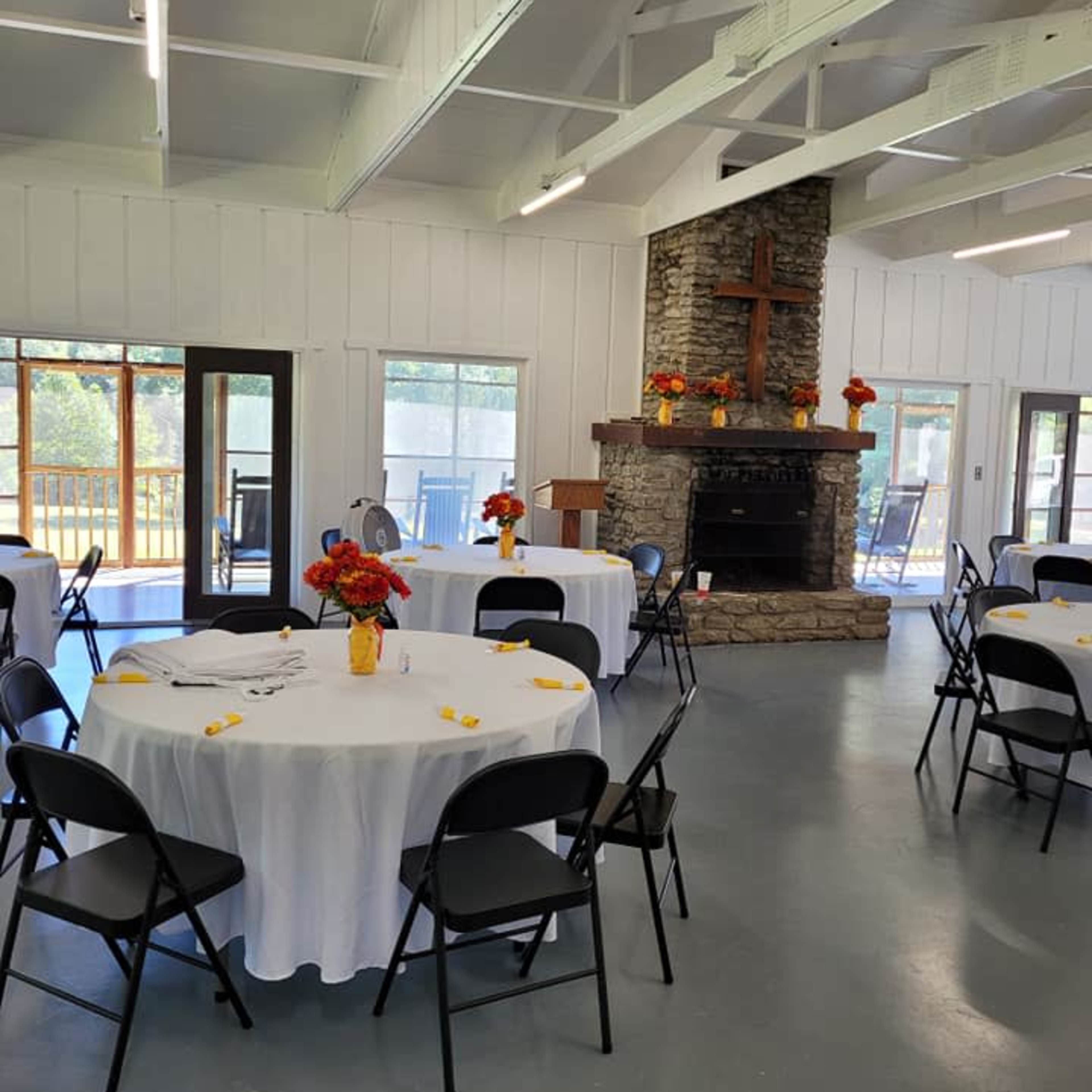 A spacious indoor setting with several round tables covered in white tablecloths and decorated with flower centerpieces, alongside a stone fireplace and a wooden cross.