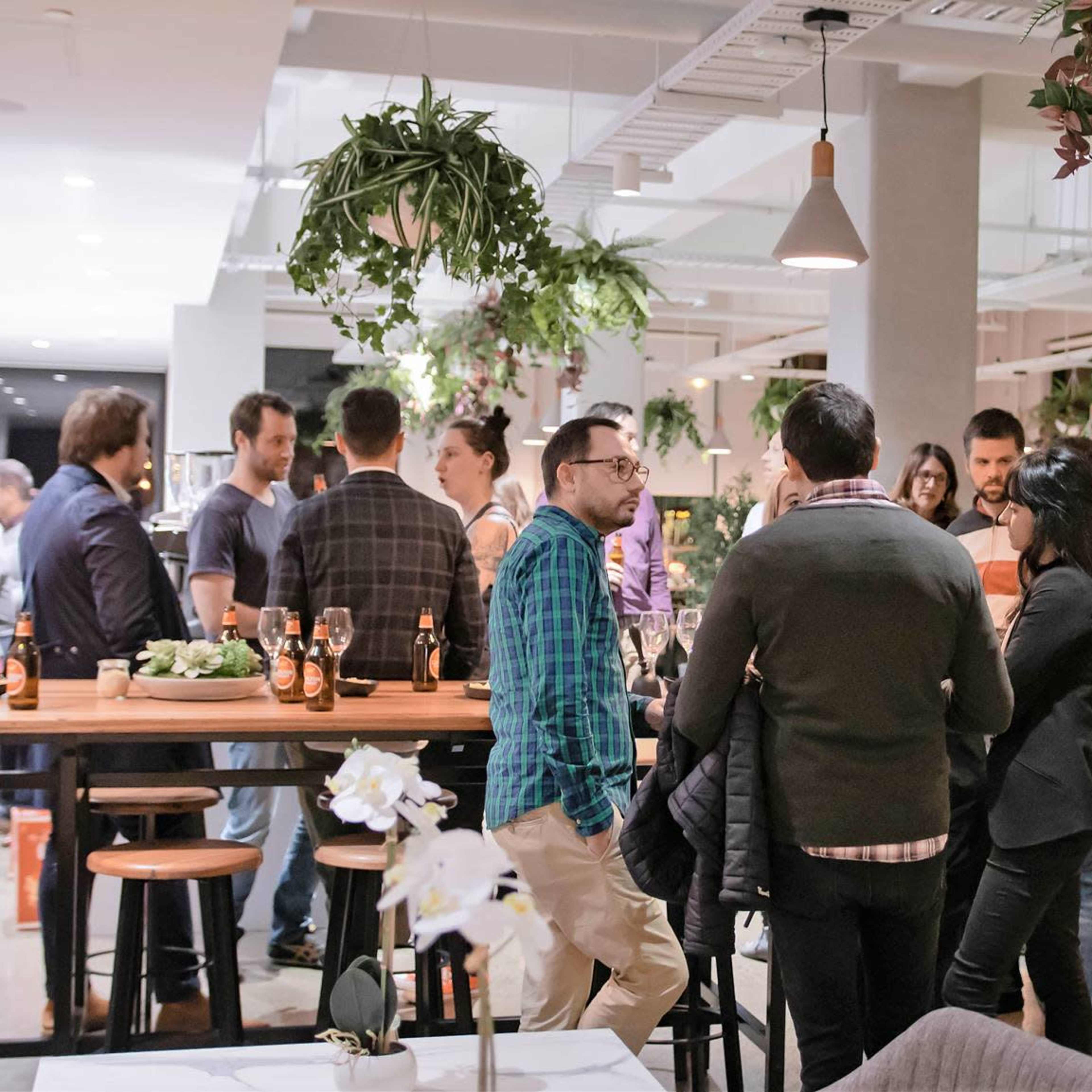 A group of people socialize in a modern indoor setting with plants hanging from the ceiling and a bar area in the background.