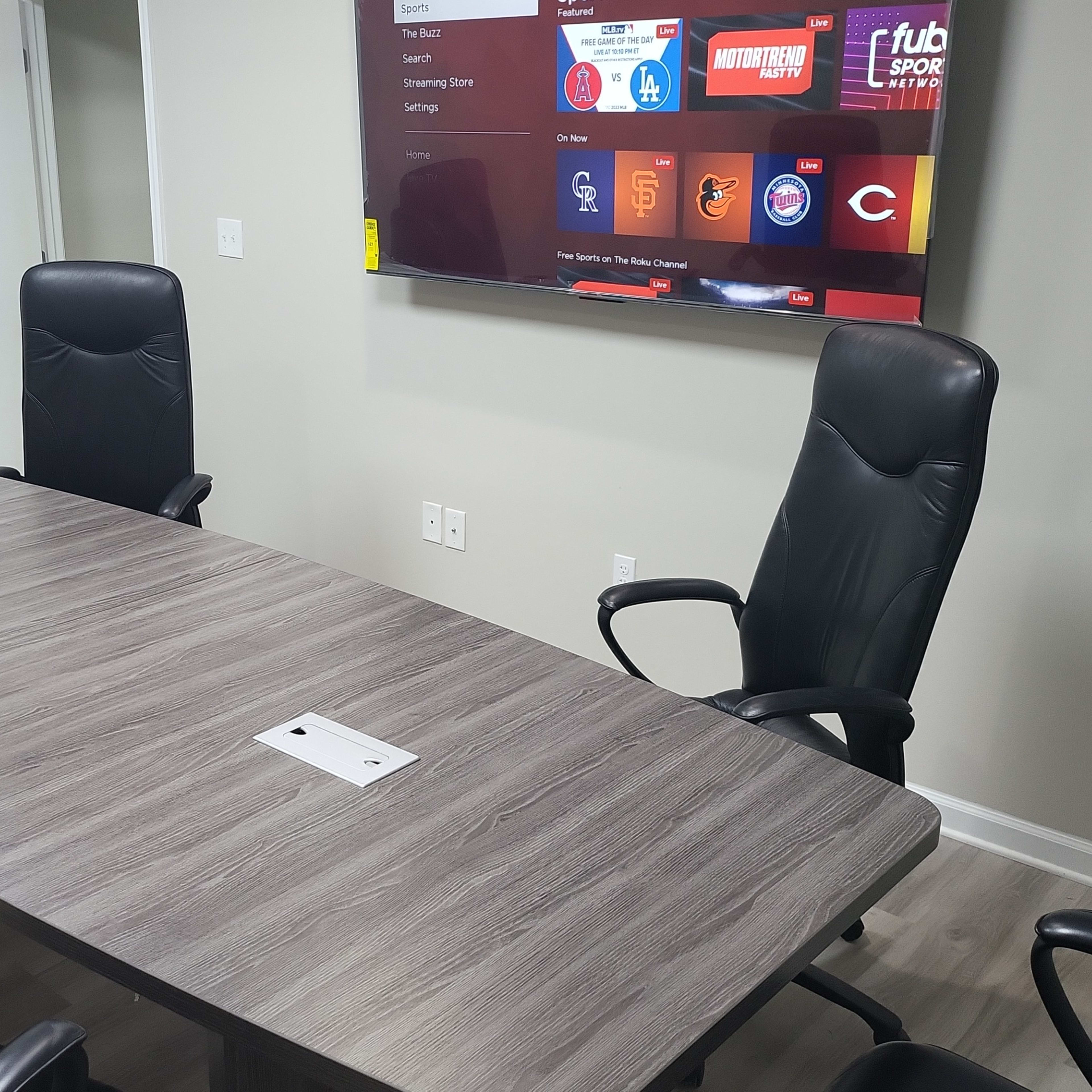 A conference room features a long wooden table surrounded by four black chairs, with a television mounted on the wall displaying various streaming options.