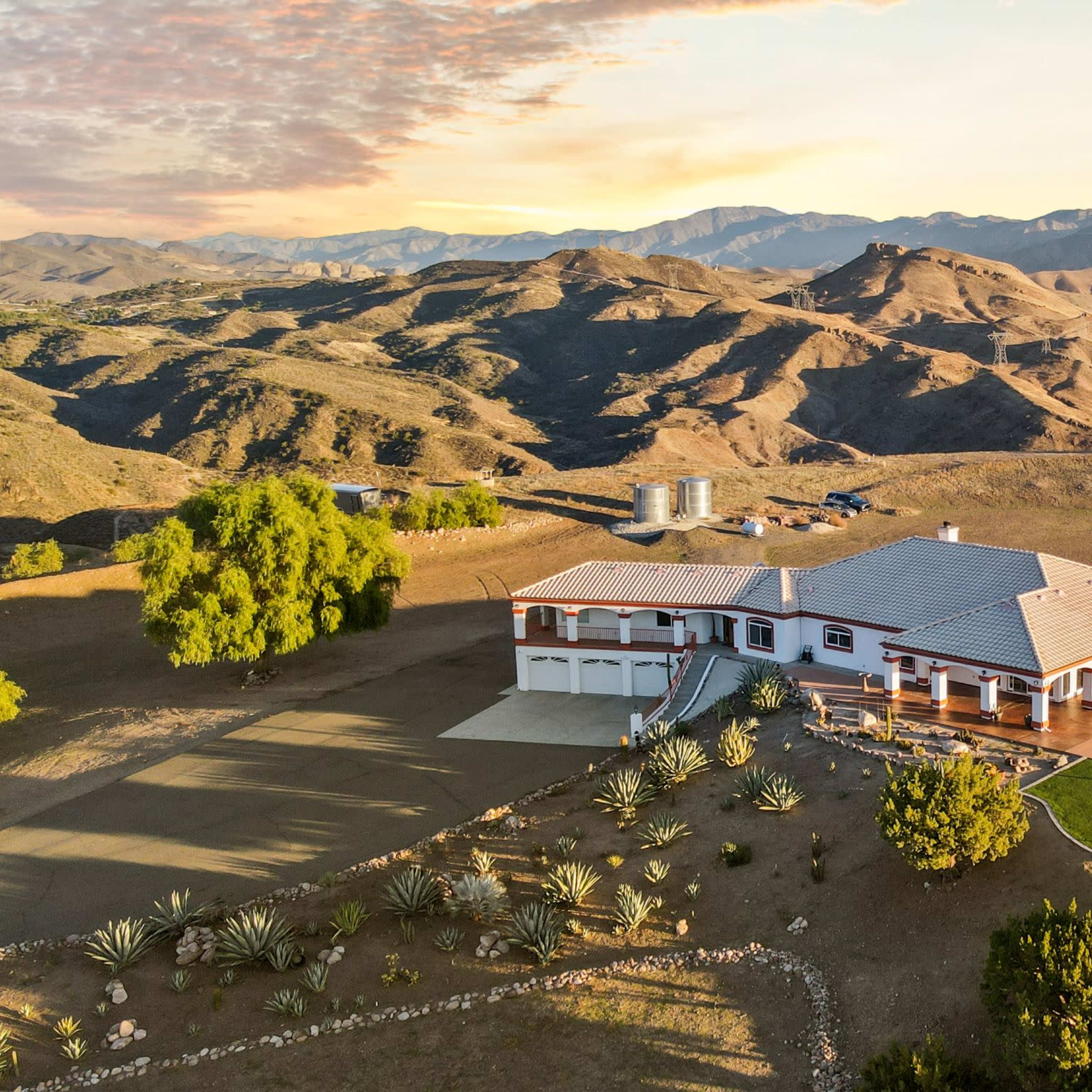 A large, single-story home surrounded by rolling hills and sparse vegetation, with a golf putting green and a parking area visible.