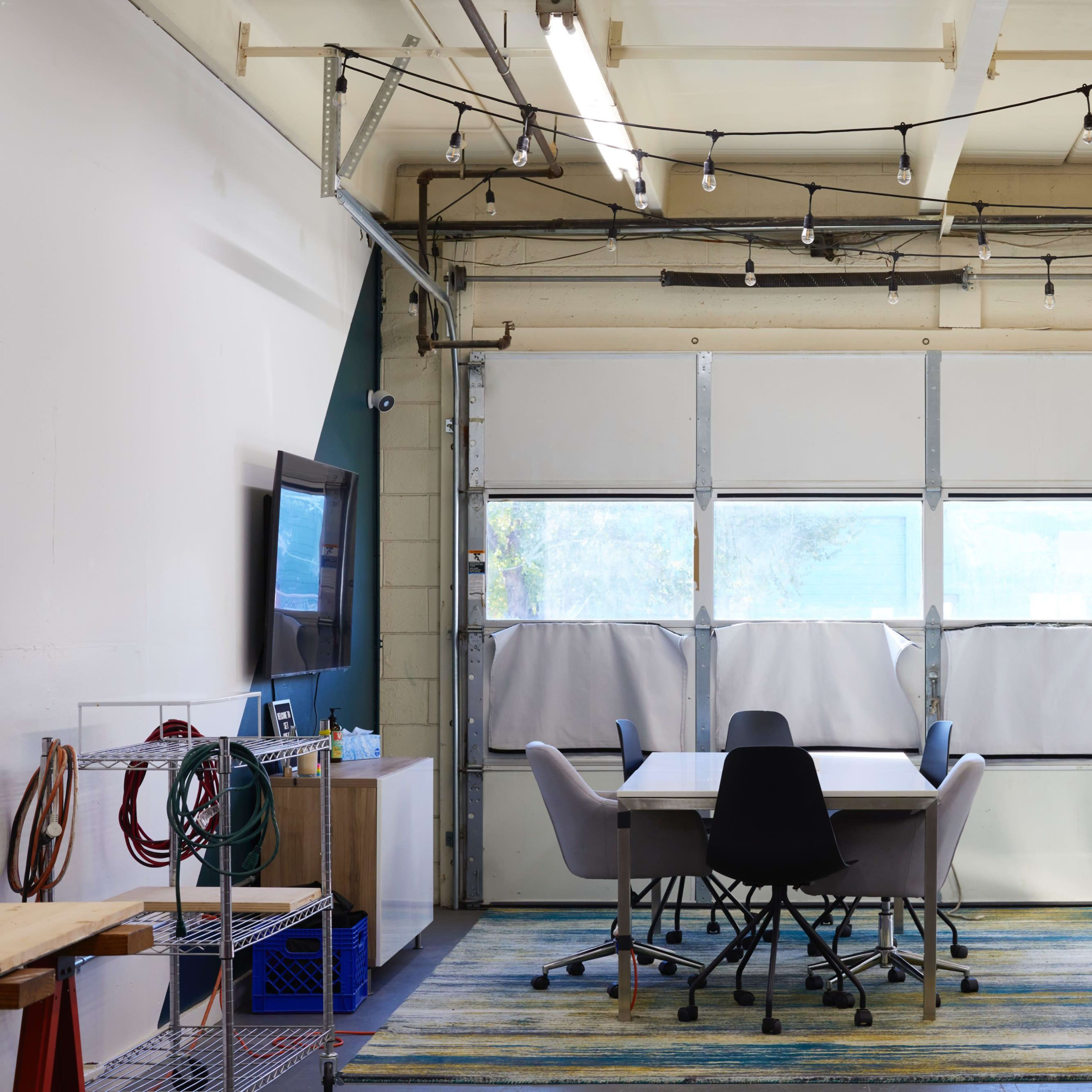 A workspace with a large table surrounded by chairs, located in a garage-like setting with large windows and exposed electrical features.