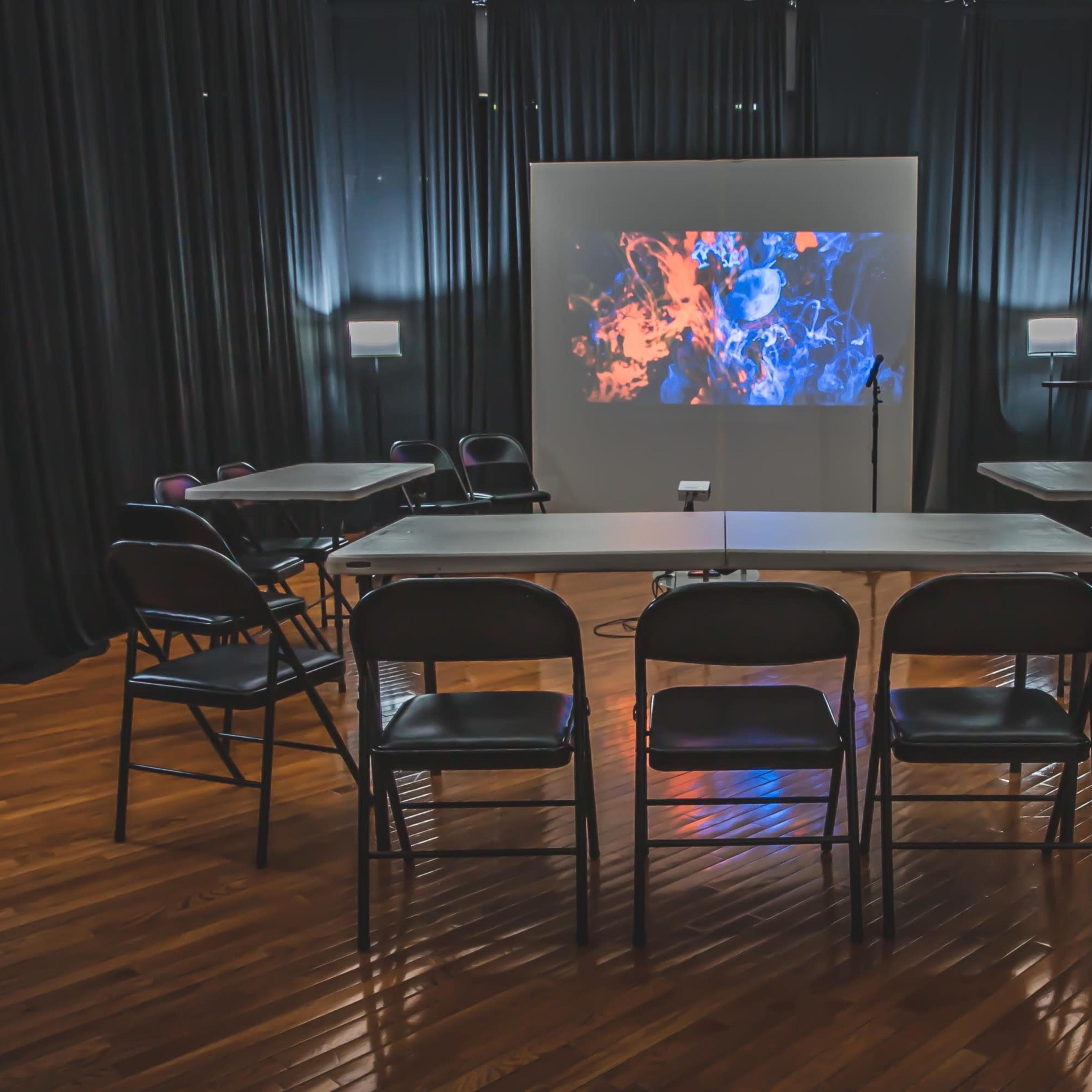 A modern meeting room features a central table surrounded by chairs, with a large screen displaying colorful visuals against a backdrop of black curtains.