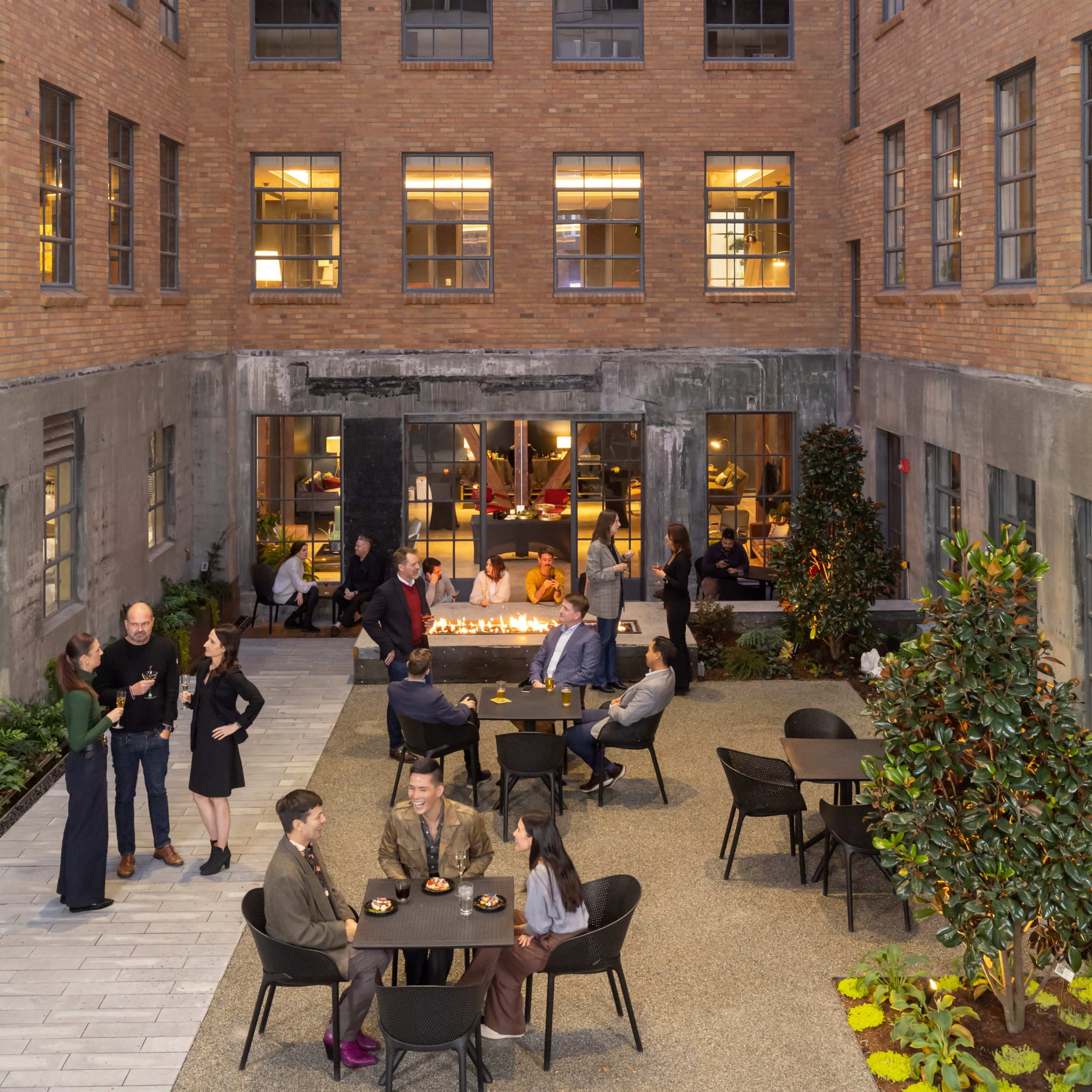 A group of people socializes around tables in a courtyard surrounded by a brick building, with a fire feature and modern lighting.
