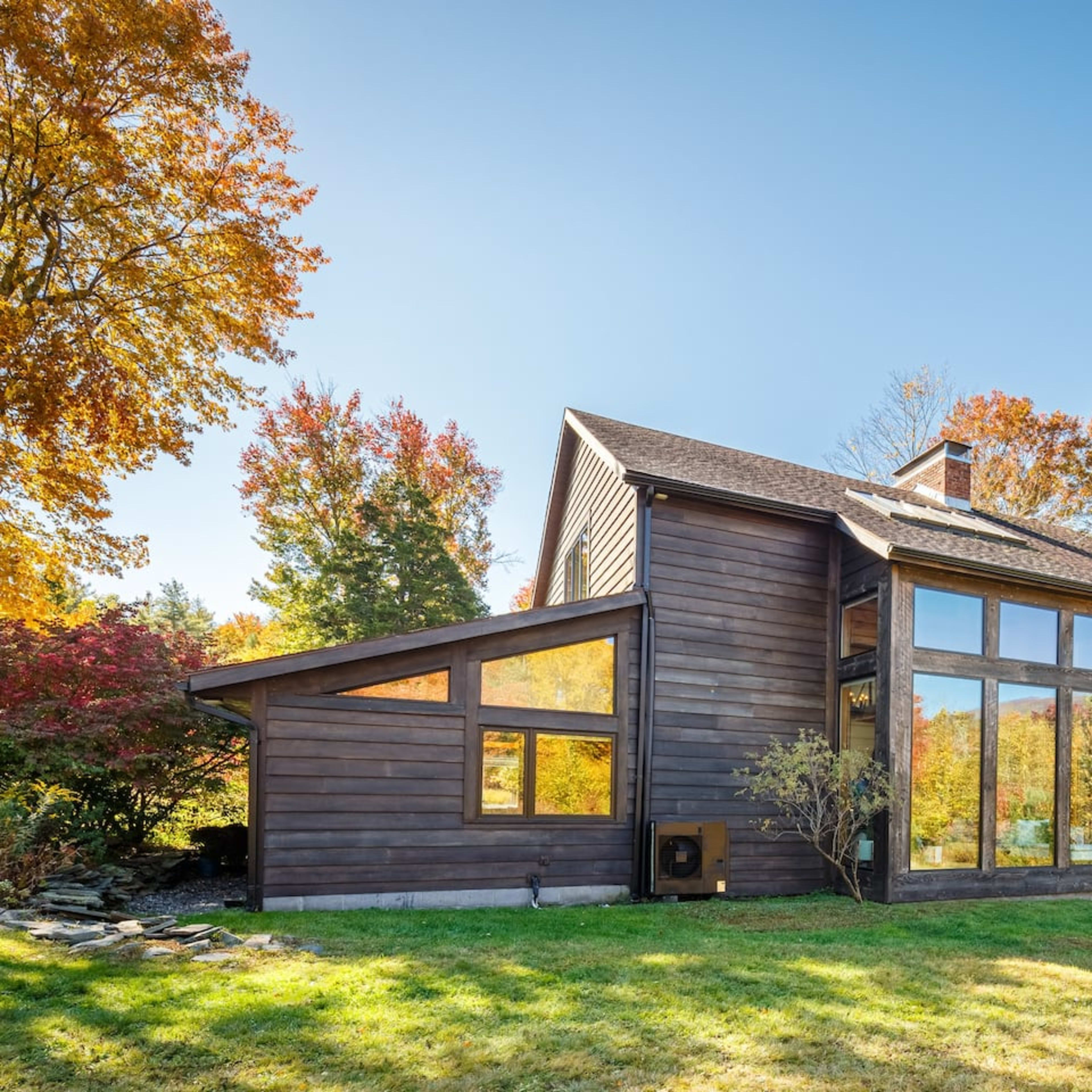 A modern wooden house with large windows is surrounded by colorful autumn trees and a clear blue sky.