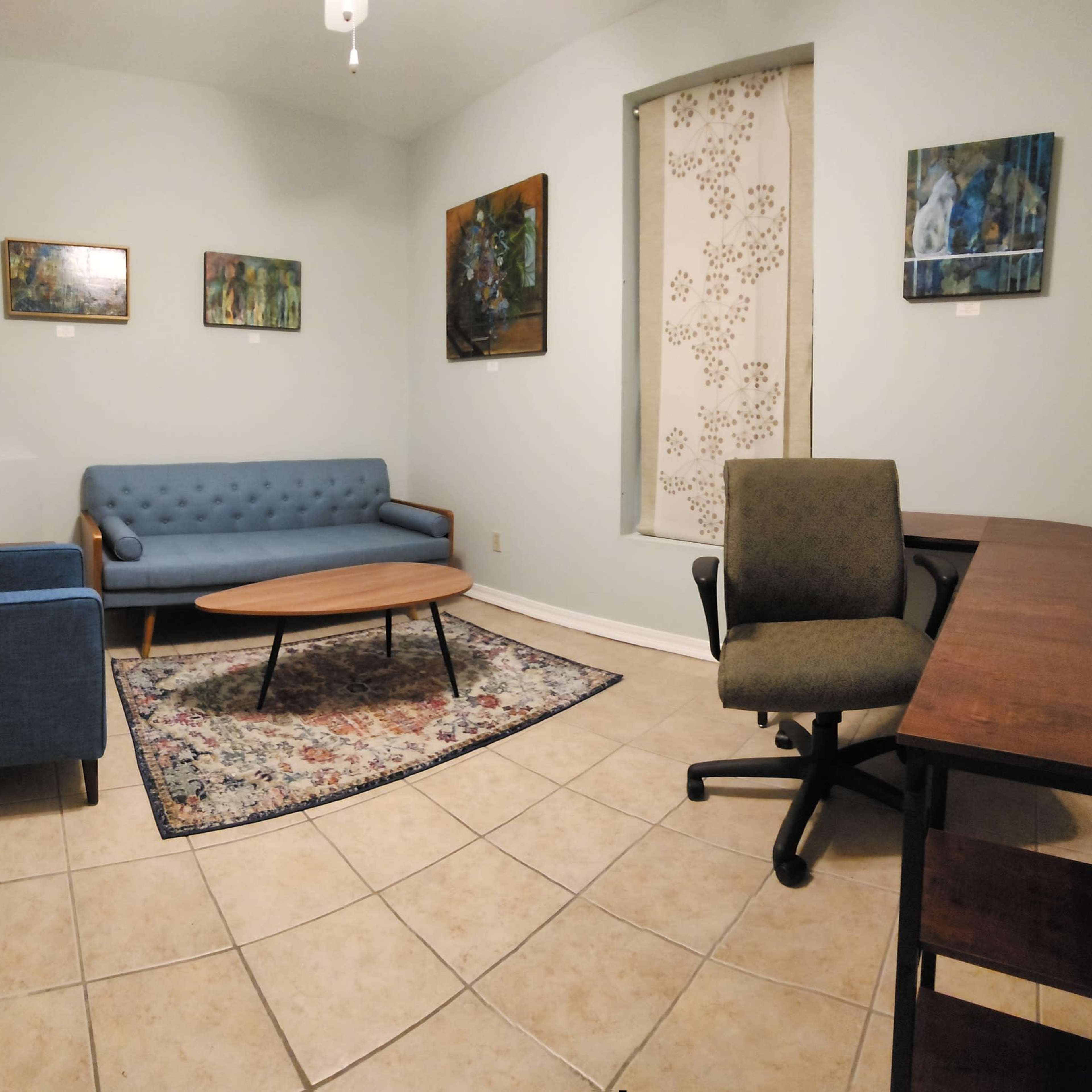 A small, softly lit room with two blue chairs, a round wooden table, and a rug, decorated with several paintings on the walls.
