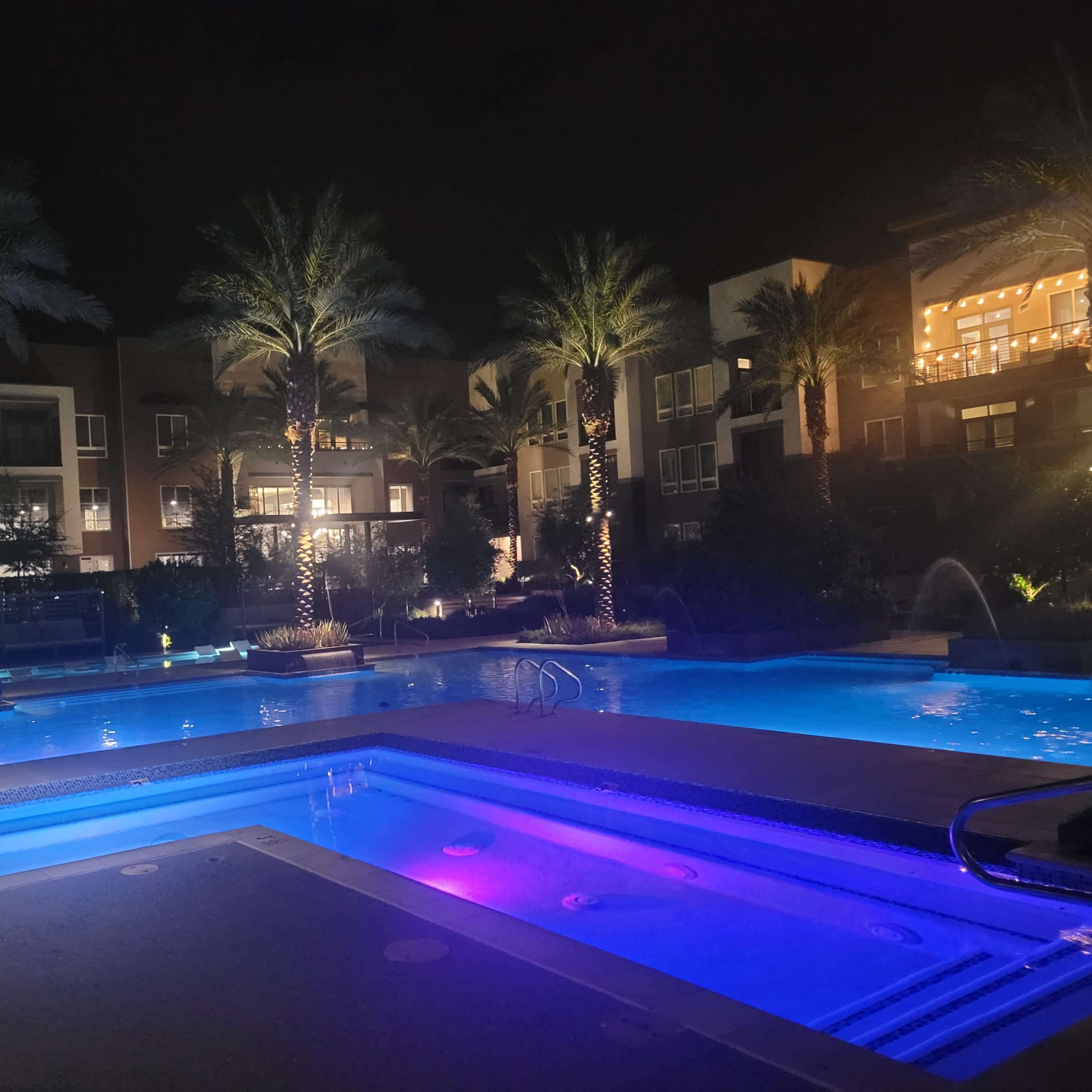 The scene depicts a brightly lit swimming pool area surrounded by palm trees and modern apartment buildings at night.