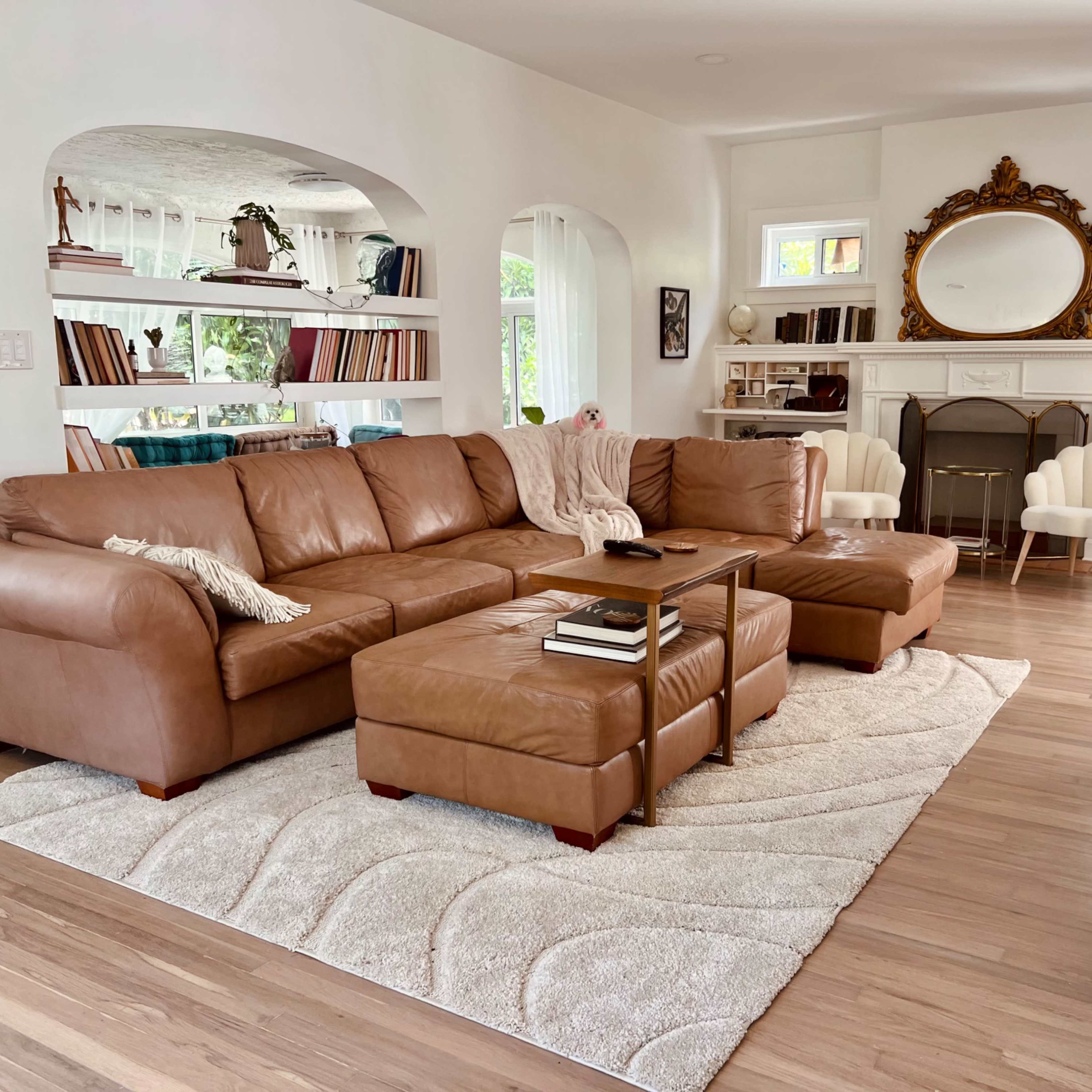 A spacious living room with a large brown leather sectional sofa, a light-colored area rug, and a wooden coffee table in front of the sofa, along with a shelf filled with books in the background.