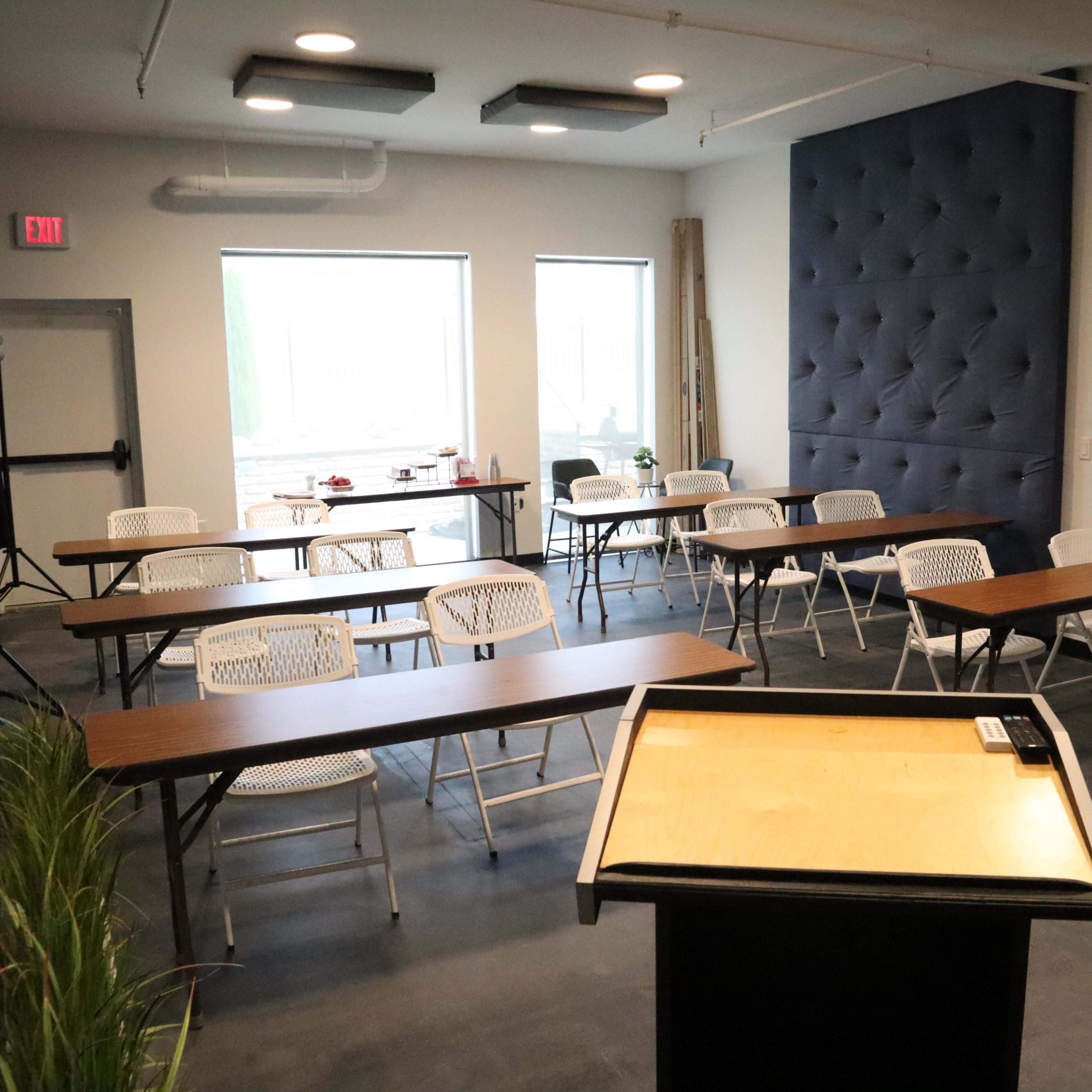 A classroom setup features several rows of tables and chairs, with a podium at the front and large windows providing natural light.
