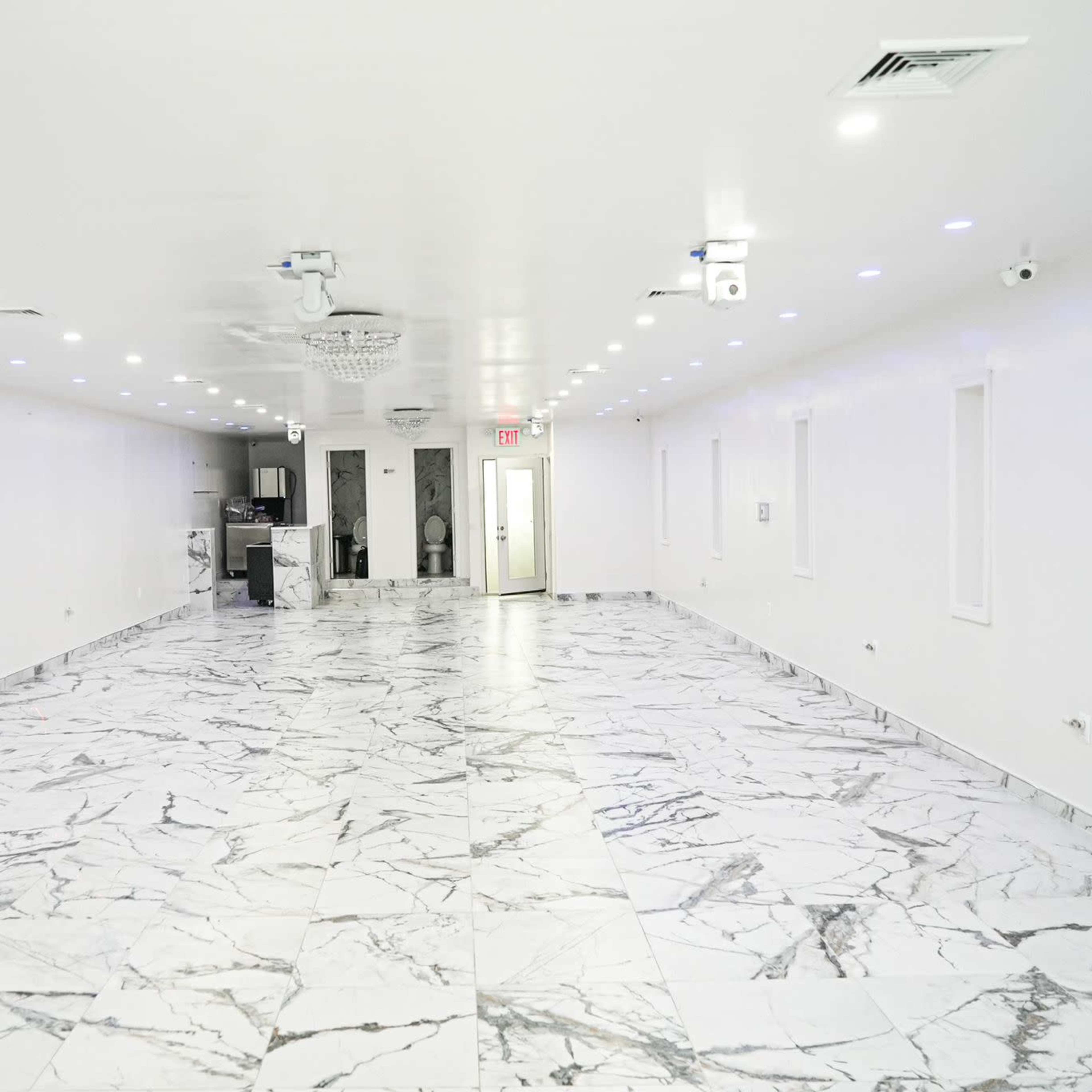 A spacious room with white walls and a marble-patterned floor, illuminated by overhead lights.