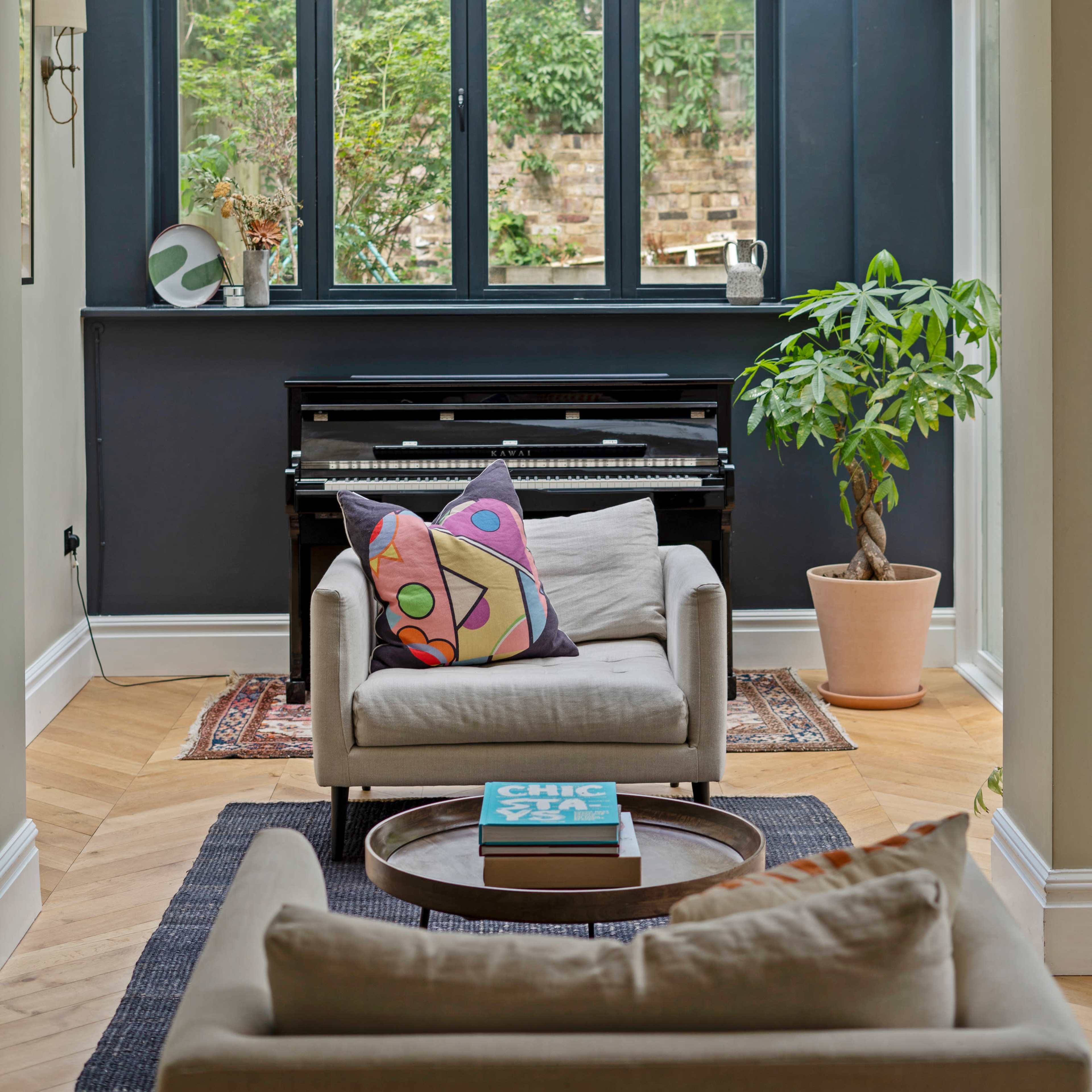 A light-filled room features a piano beneath large windows overlooking greenery, with a cozy seating area and decorative pillows on a sofa.