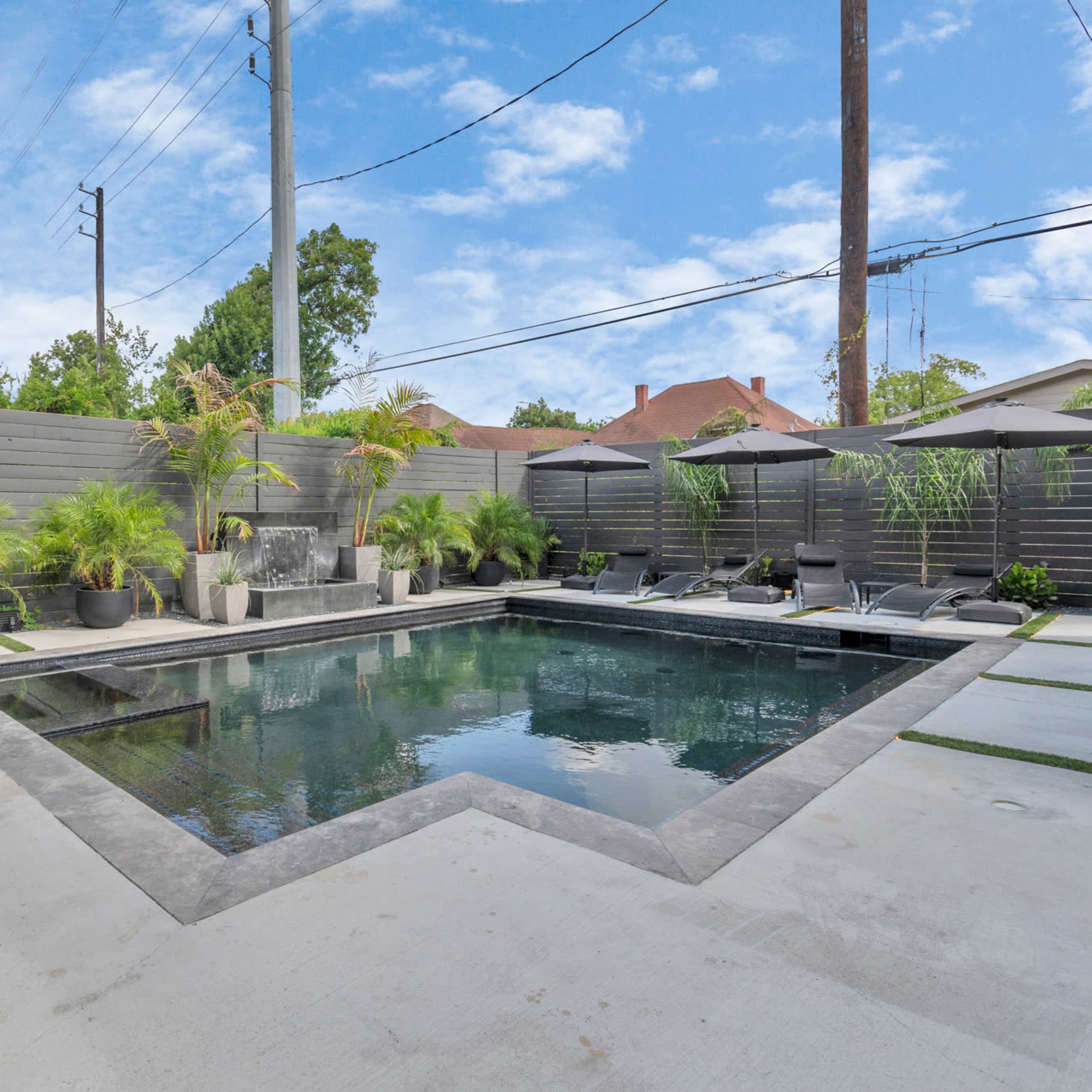 A rectangular backyard pool surrounded by concrete and landscaped with potted plants and lounge chairs under umbrellas.