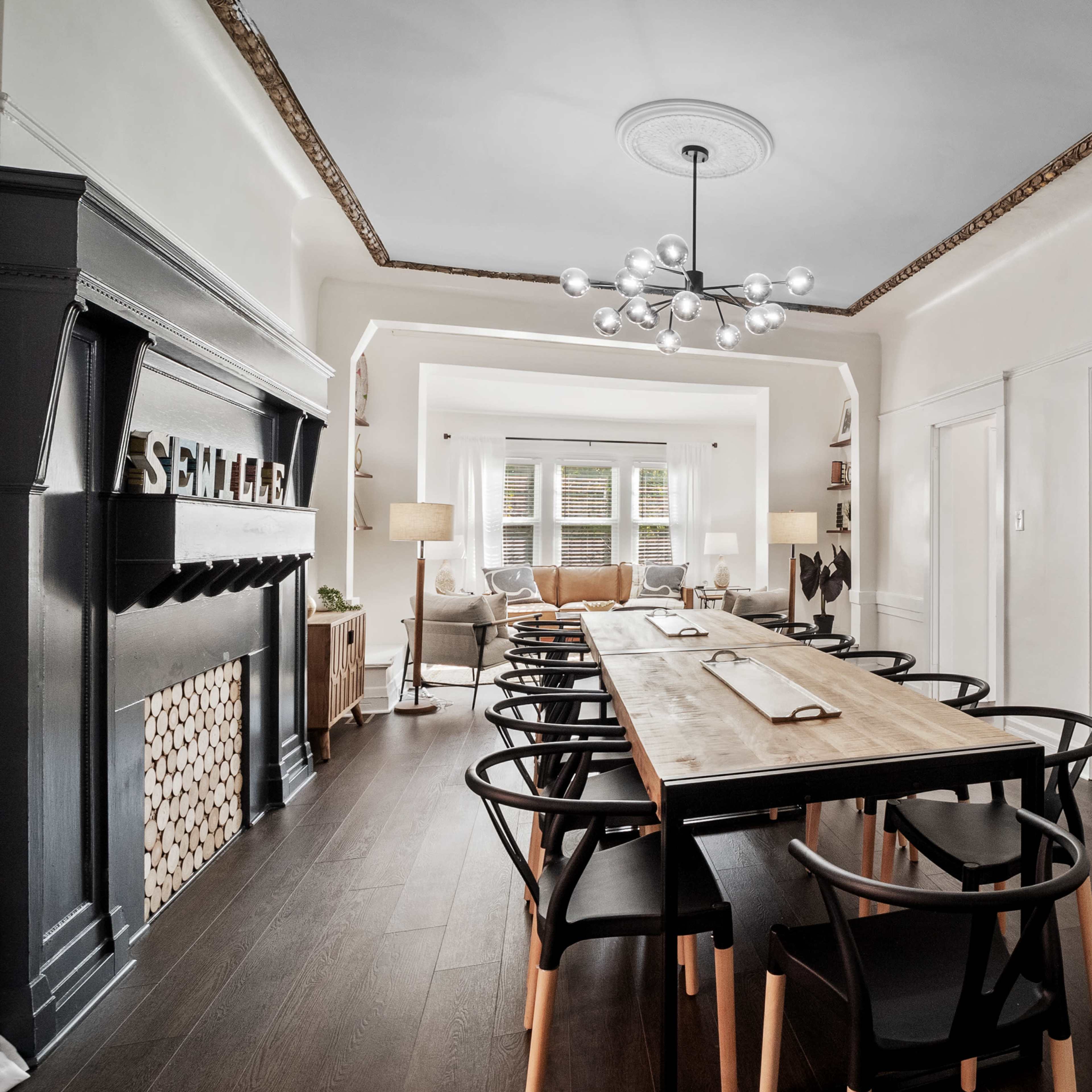 A long wooden dining table with black chairs is set in a well-lit room featuring a decorative fireplace and framed artworks on the walls.