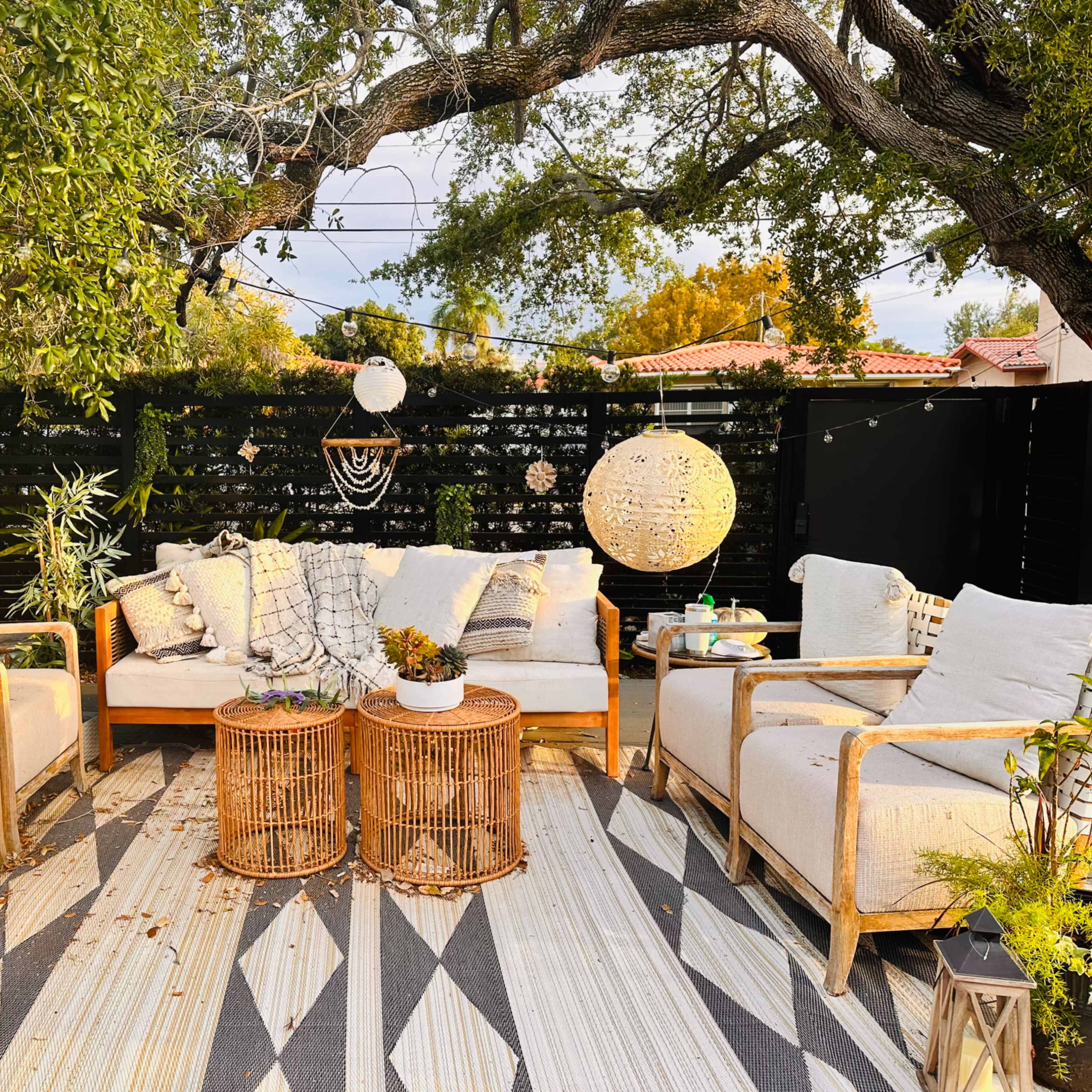 A cozy outdoor seating area featuring light-colored sofas and chairs arranged around woven coffee tables, with greenery and hanging decorations overhead.
