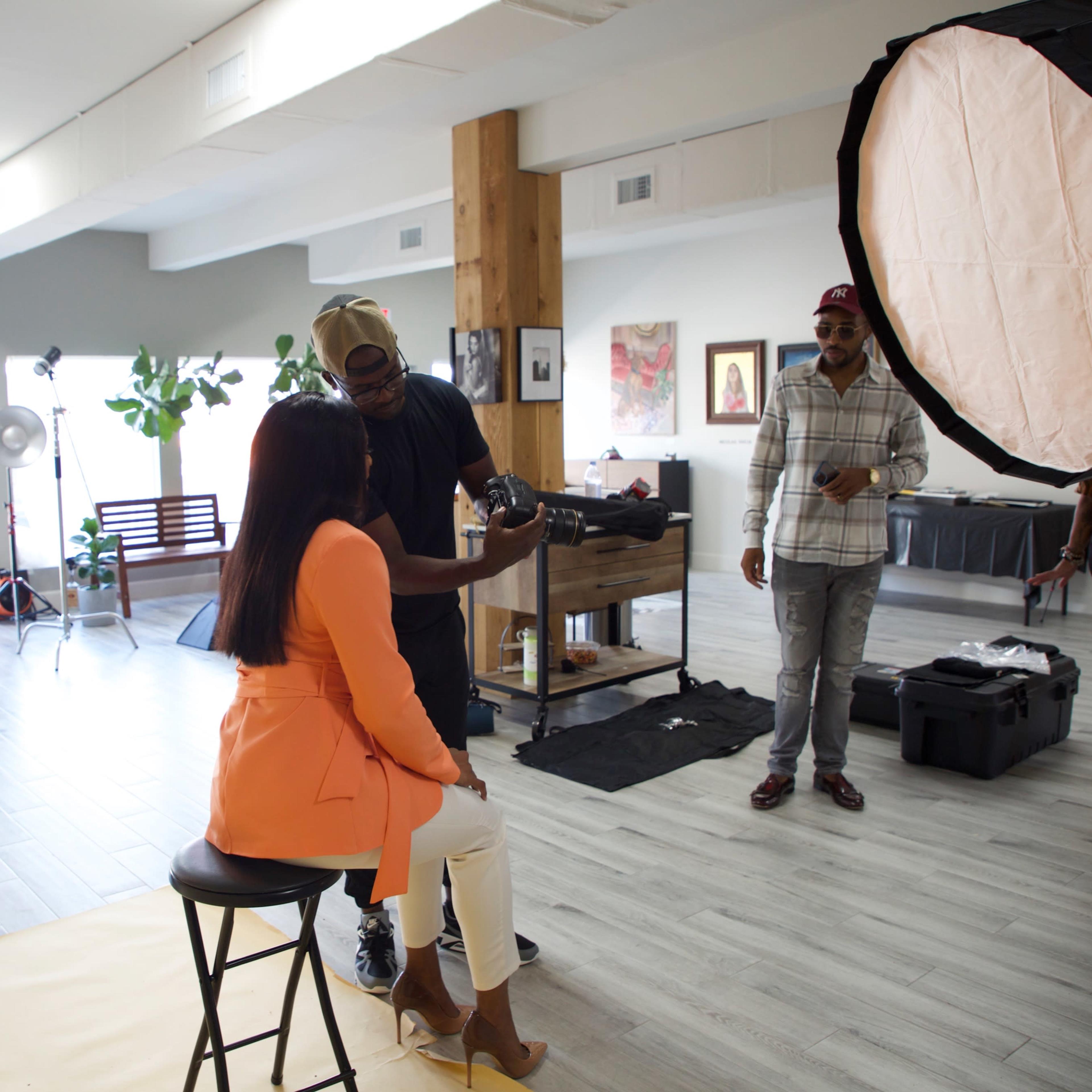 A photographer prepares a model for a photoshoot in a well-lit studio setting with various lighting equipment and props.