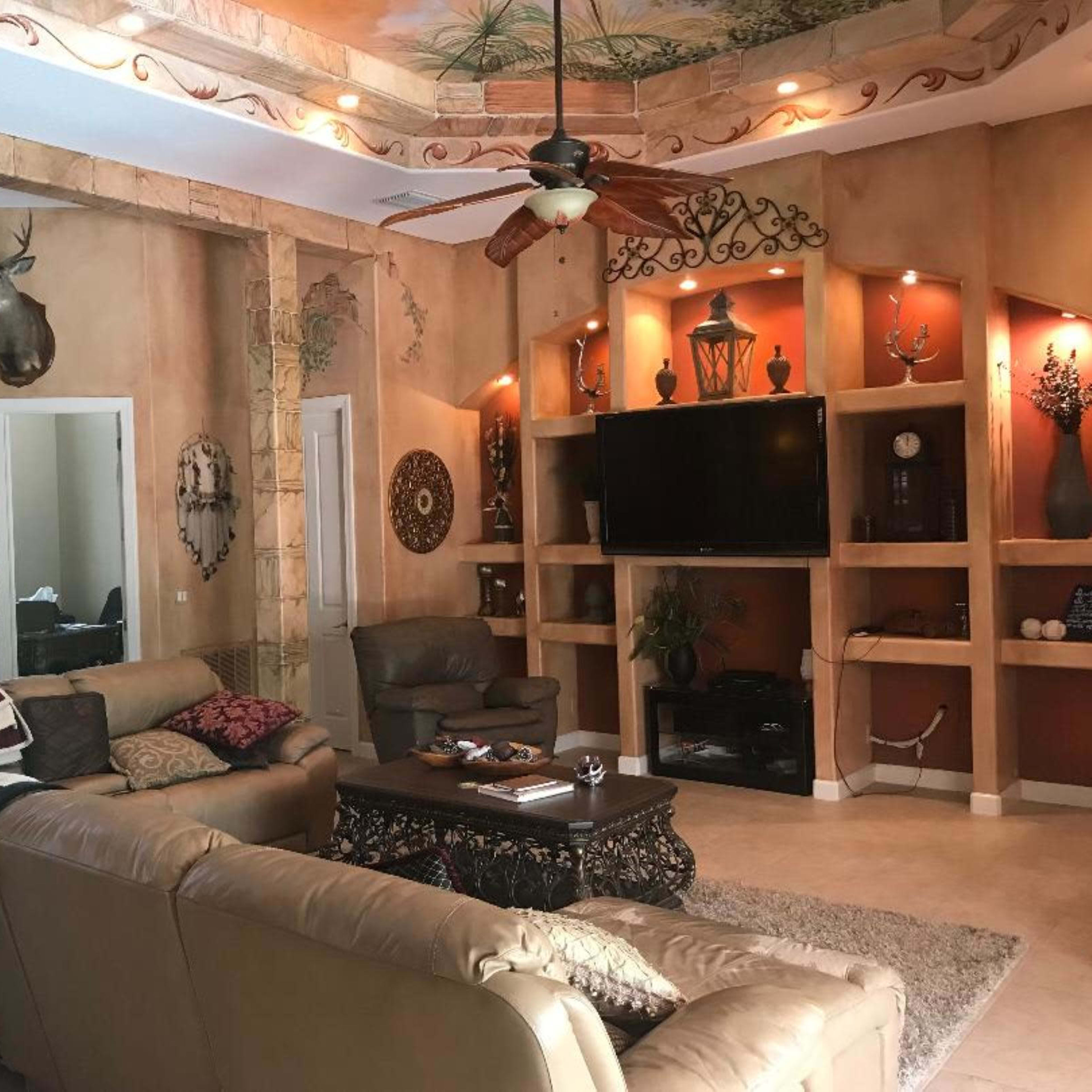 A living room features a large sectional sofa, a television mounted on a wall unit, and decorative elements including a mounted deer head and various wall accents.