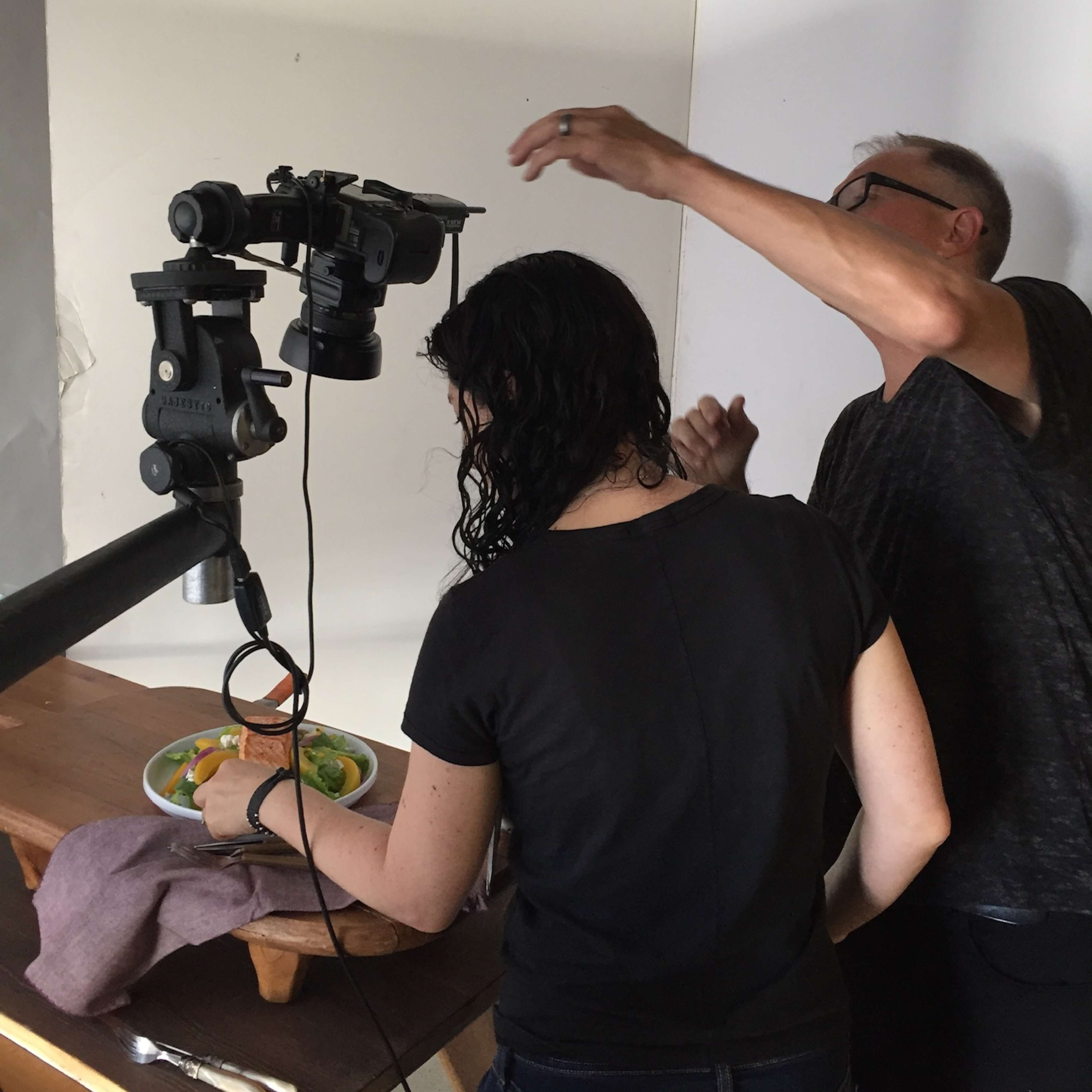 A person adjusts a camera setup while another person arranges food on a table for a photo shoot.