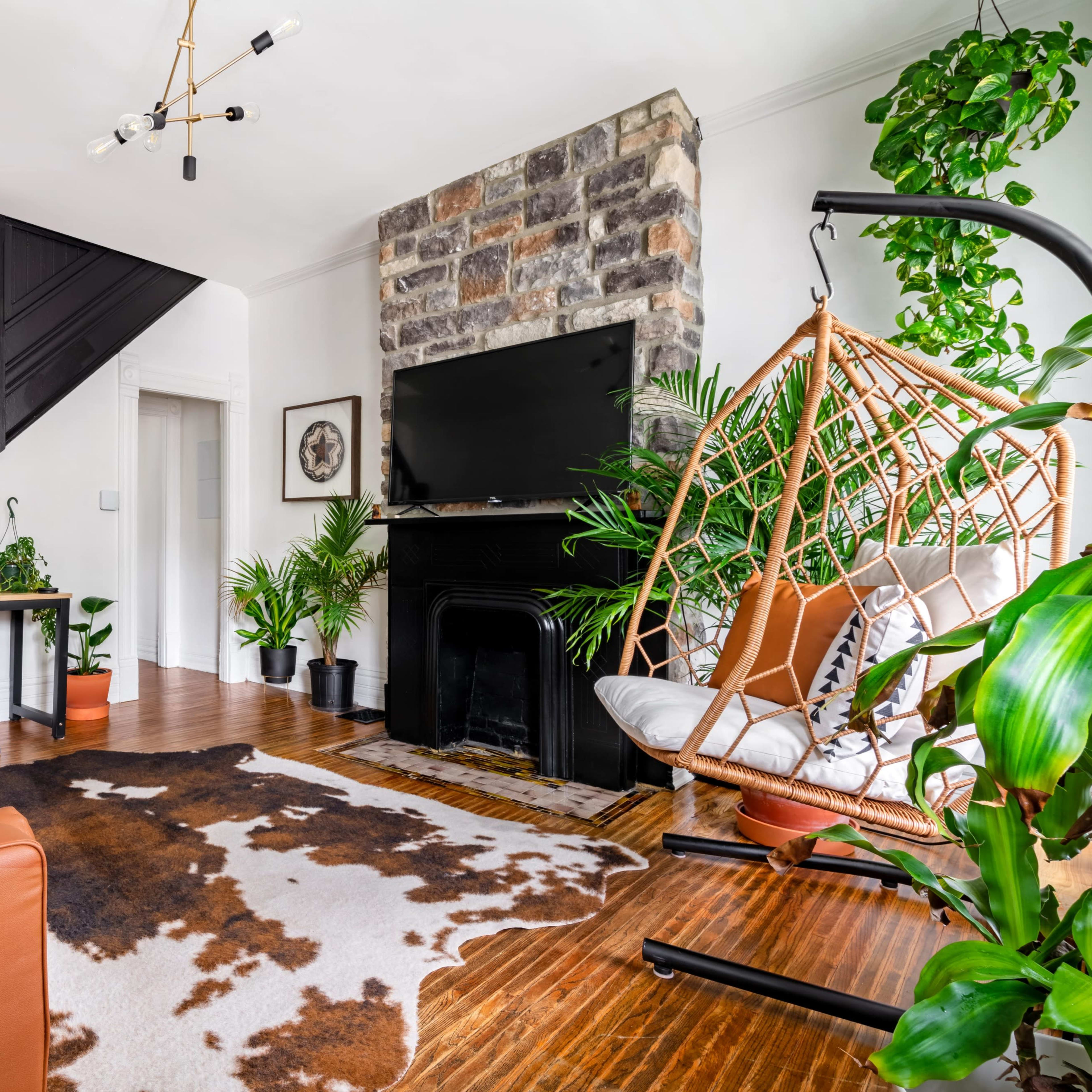 The room features a cozy living area with a brown leather sofa, a cowhide rug, plants throughout, and a large television on a stone fireplace wall.