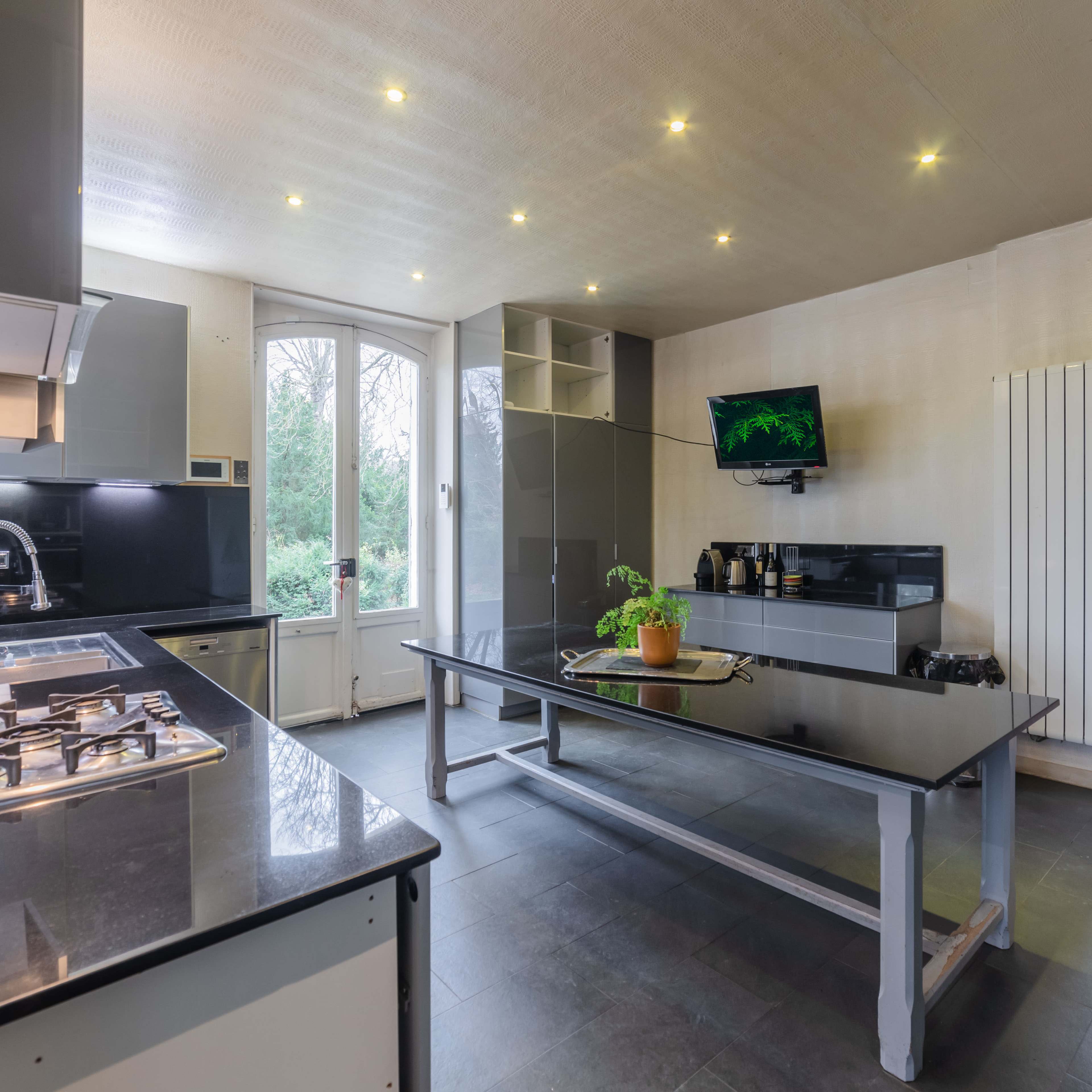 A modern kitchen features a central black table, sleek cabinetry, a gas stove, and a television mounted on the wall, with large windows that offer a view of the outdoors.