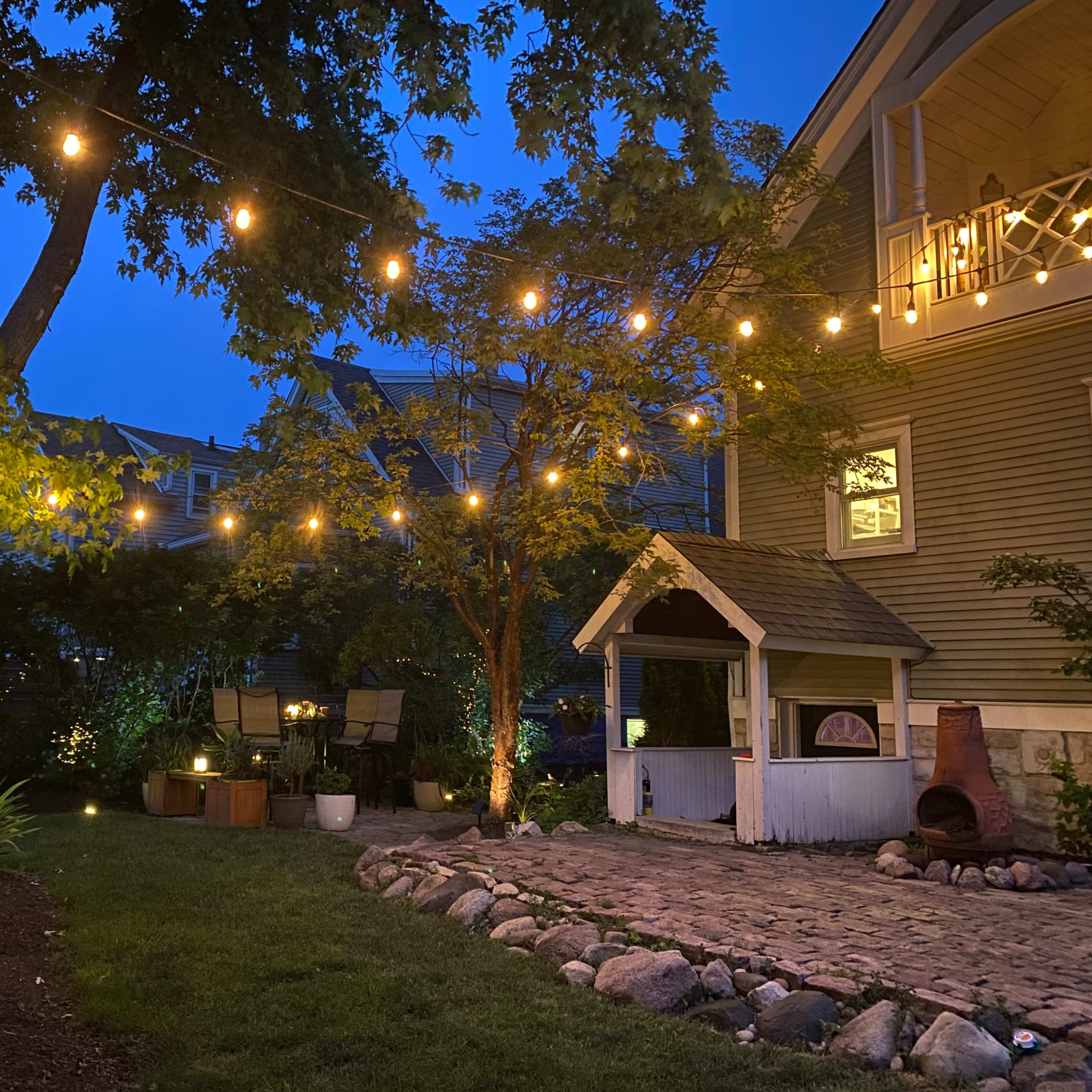 The backyard features string lights illuminating a small playhouse, surrounded by greenery and a stone path under a twilight sky.