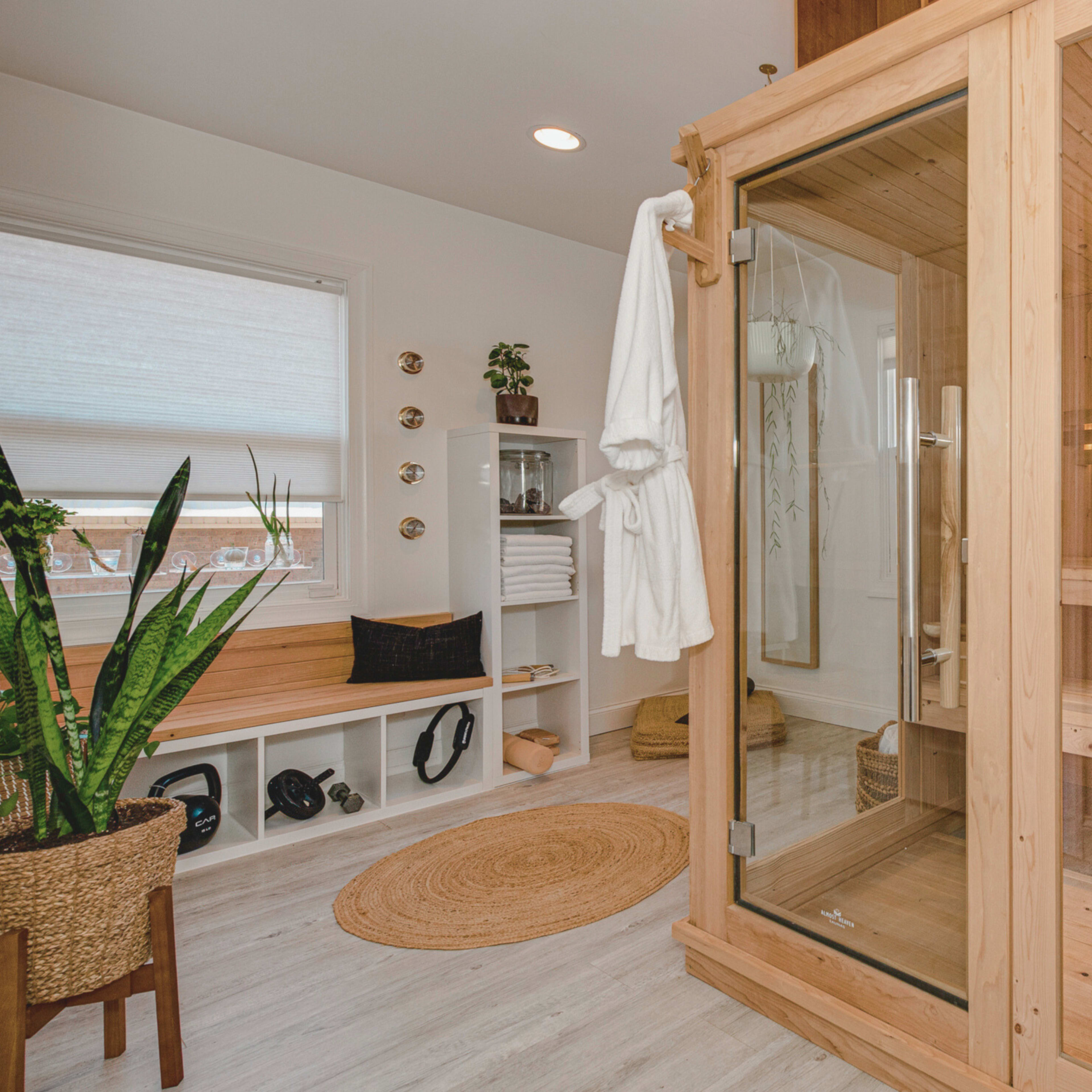 A modern bathroom featuring a wooden sauna, a plant, a white robe hanging on the door, and organized shelves with towels.