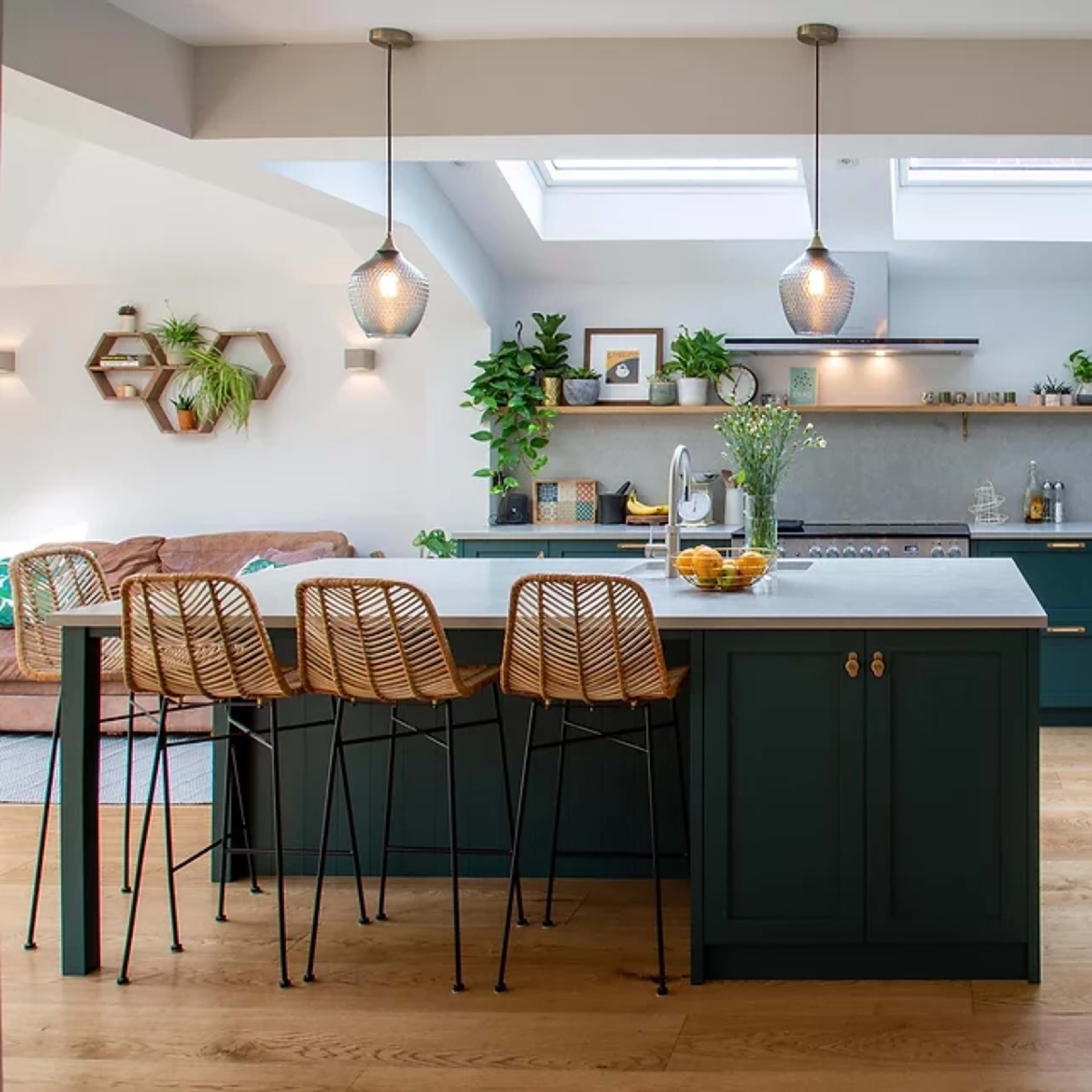 A modern kitchen with a central island, bar stools, and skylights, featuring green cabinetry and plants.