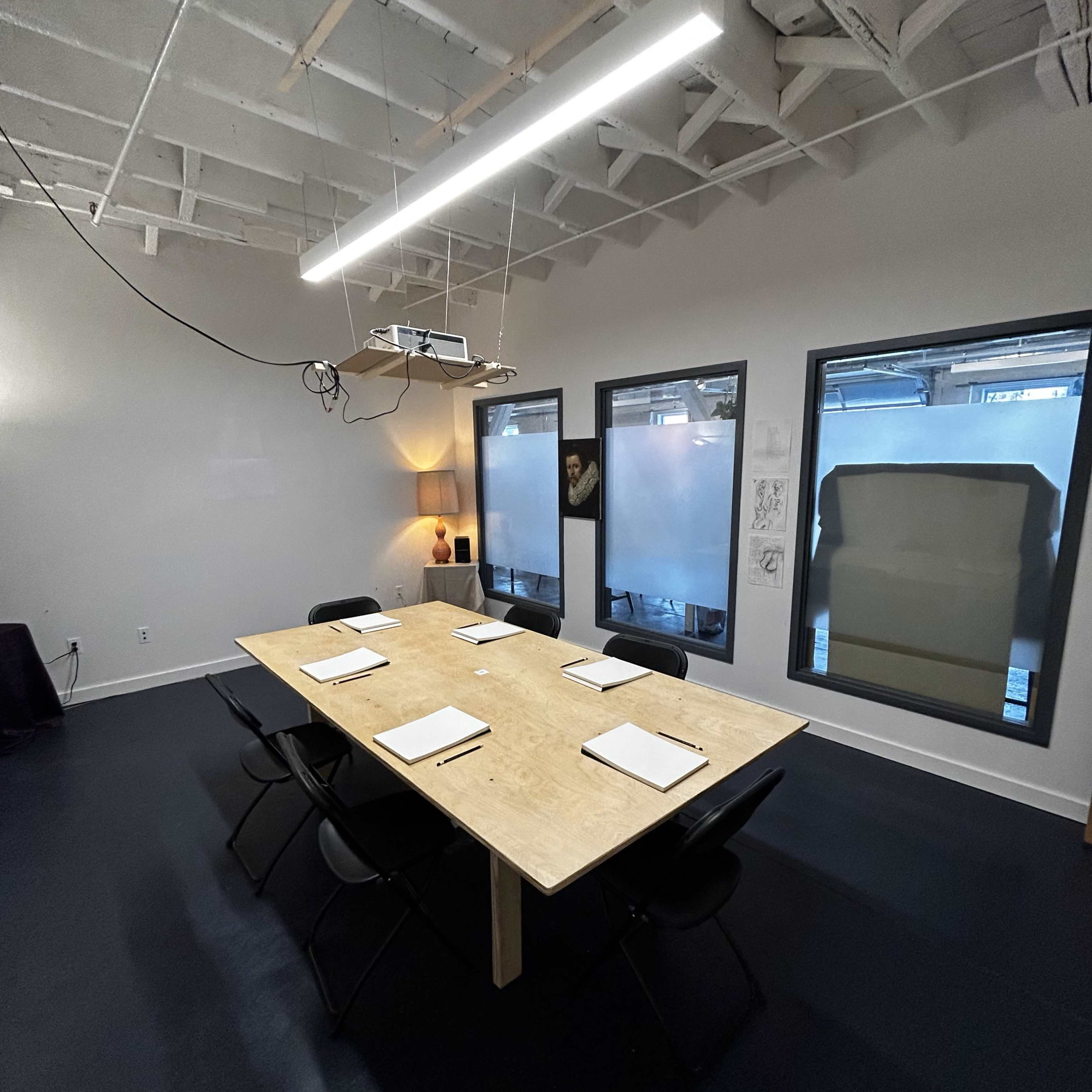 A long wooden table with chairs is arranged in a modern conference room featuring a ceiling with exposed beams and large windows.