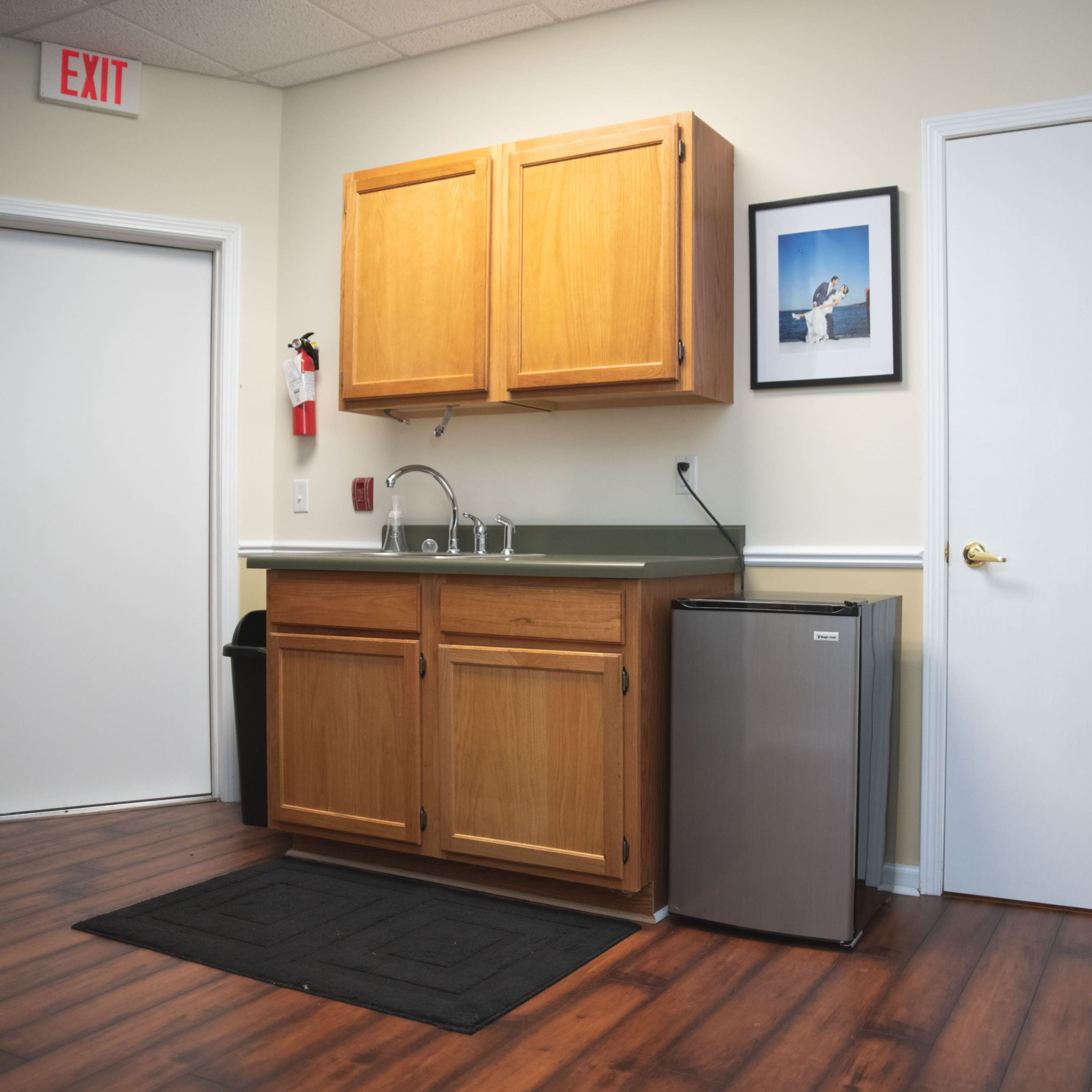 A small kitchenette area with wooden cabinets, a sink, a mini-fridge, and a door leading to another room.