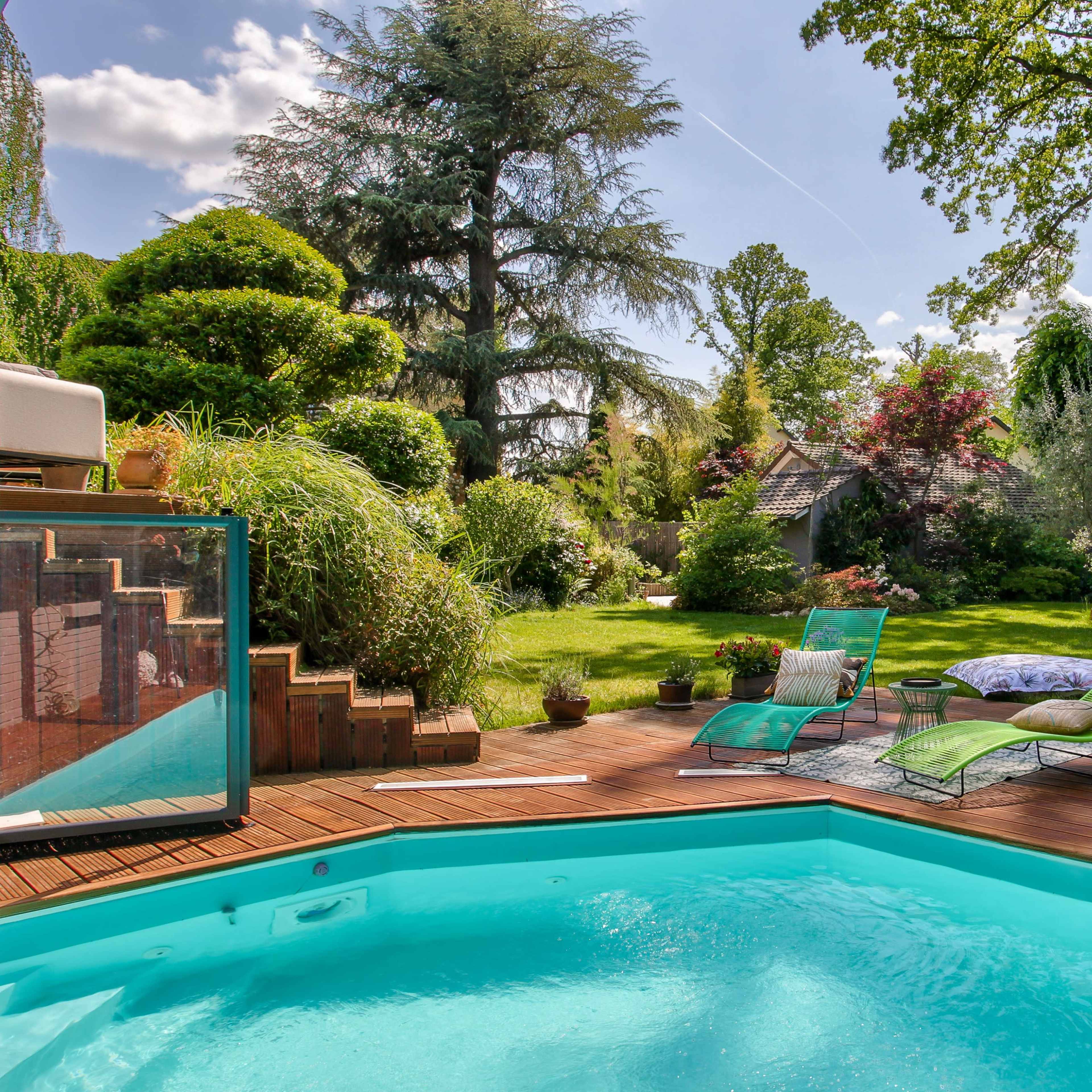 A clear blue swimming pool is bordered by a wooden deck and surrounded by a landscaped garden featuring trees, shrubs, and lounge chairs.