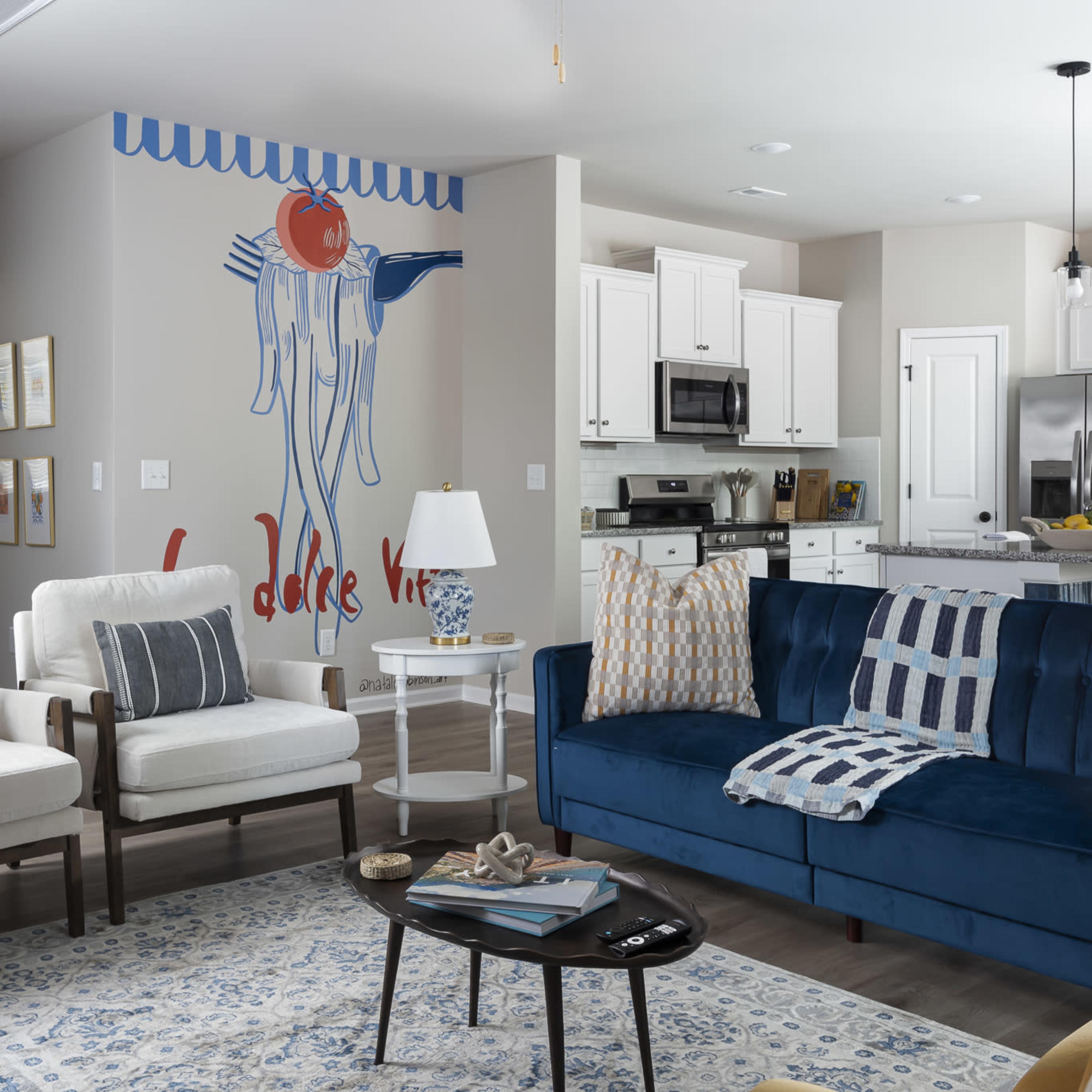A modern living room features a blue sofa, two white armchairs, and a kitchen in the background with a large wall mural of a hanging tomato.