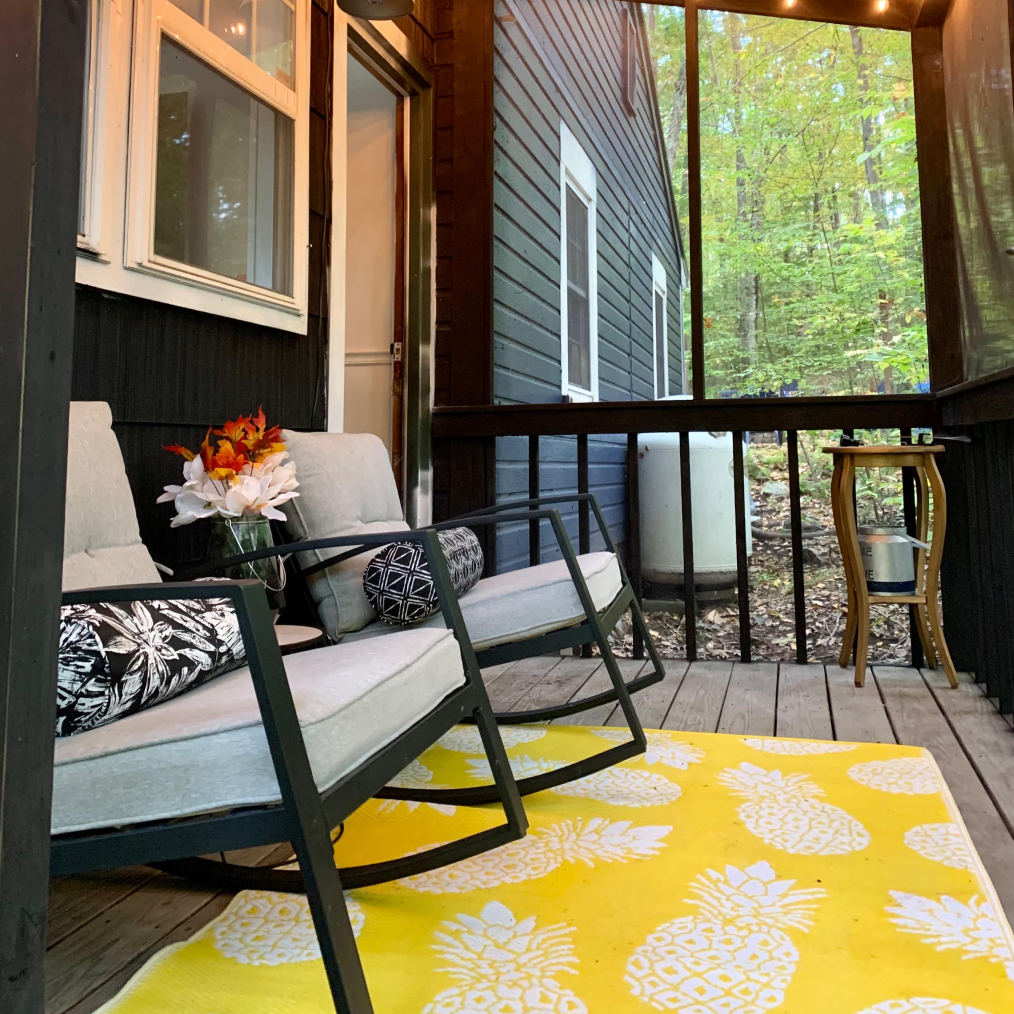A cozy porch features two chairs, a small table, and an area rug with a pineapple design, all illuminated by string lights overhead.