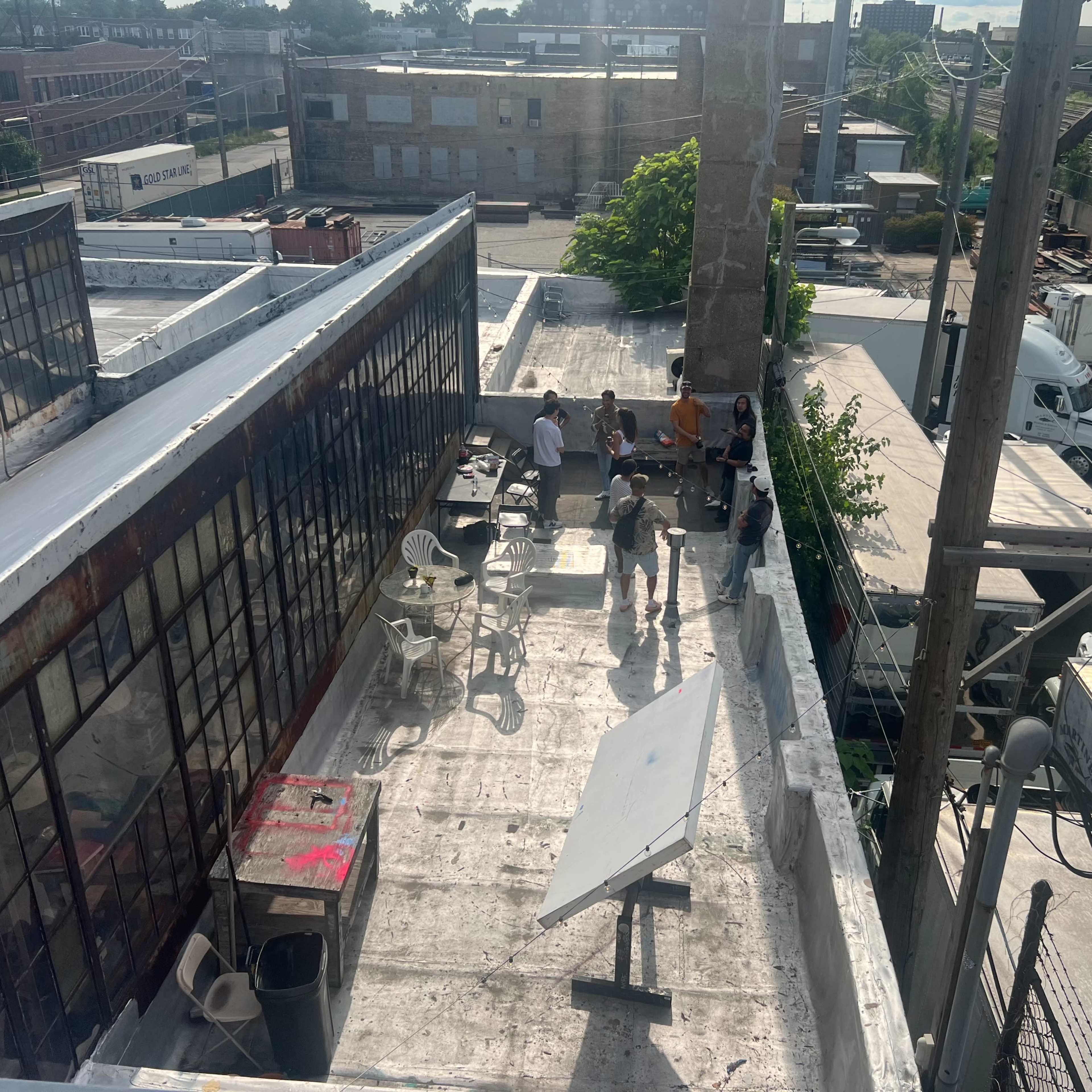 A group of people stands on a rooftop terrace surrounded by industrial buildings and parked trucks.