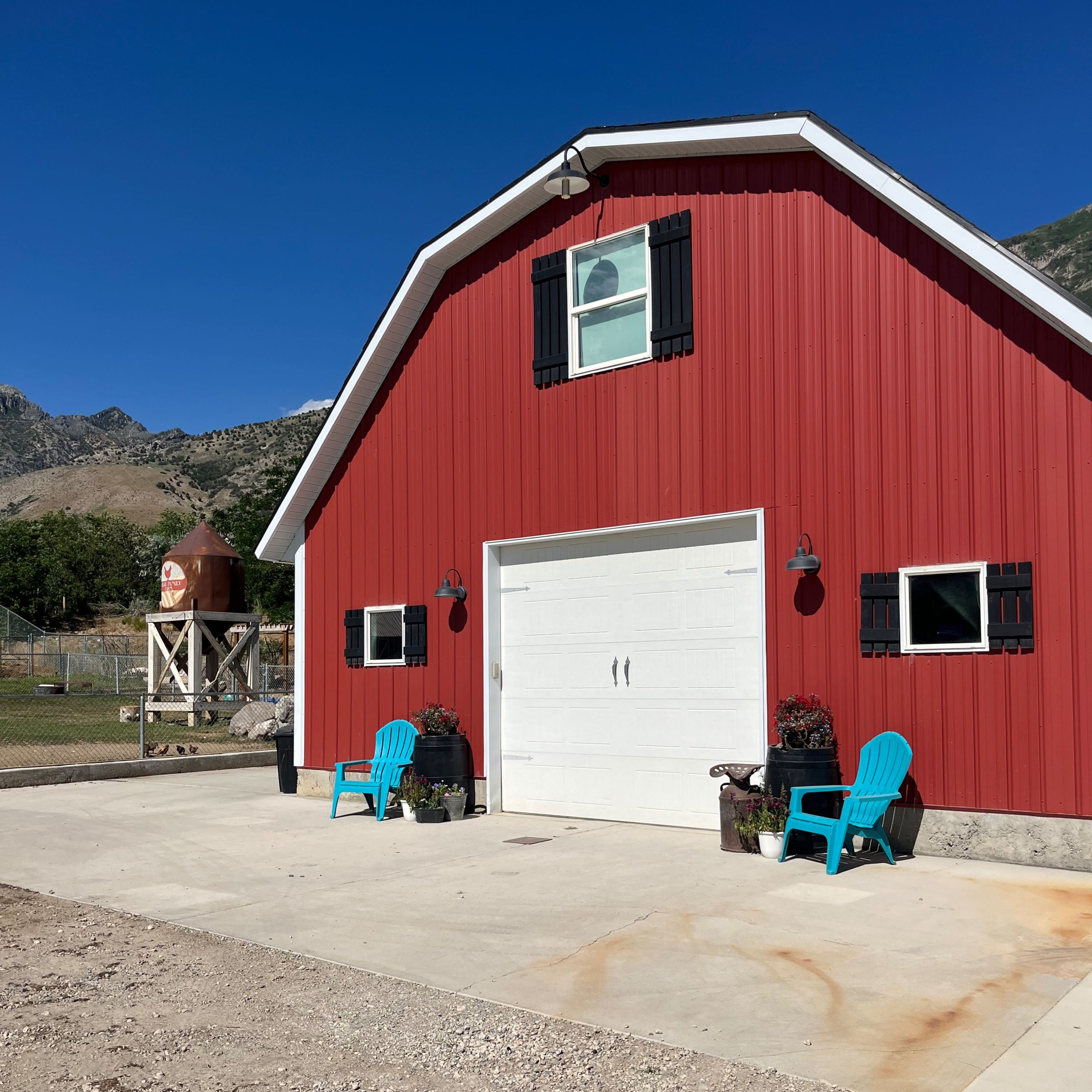 A red barn with a white garage door, two turquoise chairs in front, and a water tank nearby under a clear blue sky.