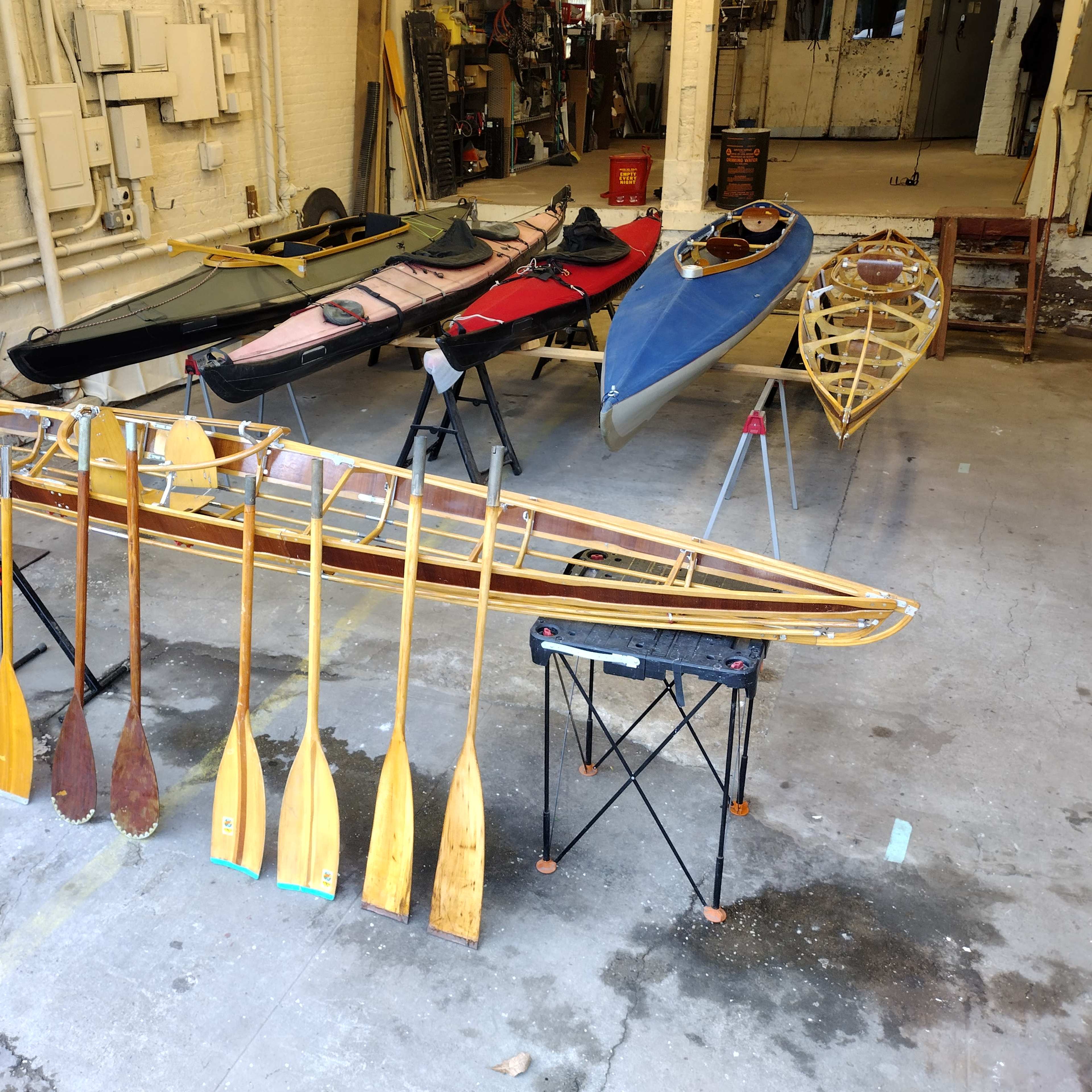 A set of kayaks and a wooden kayak frame with paddles are displayed in a spacious workshop.